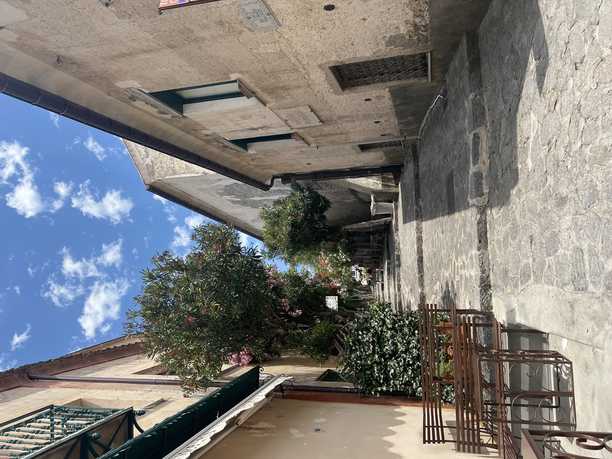 The image depicts a sun-drenched cobblestone stairway in Ravello, Italy. Two individuals, a woman and a man, are ascending the stairs, viewed from behind. To the left of the stairway, there is a building with beige walls, a green awning, and a wrought-iron balcony. Further up the stairs, a tree with pink blossoms is prominent, adding a touch of color to the scene. The architecture suggests an old European town with narrow streets and stone construction. The sky above is blue with scattered white clouds. In the foreground, to the left, metal benches and tables are arranged, indicating a potential seating area. The overall impression is one of a peaceful, picturesque Italian street.