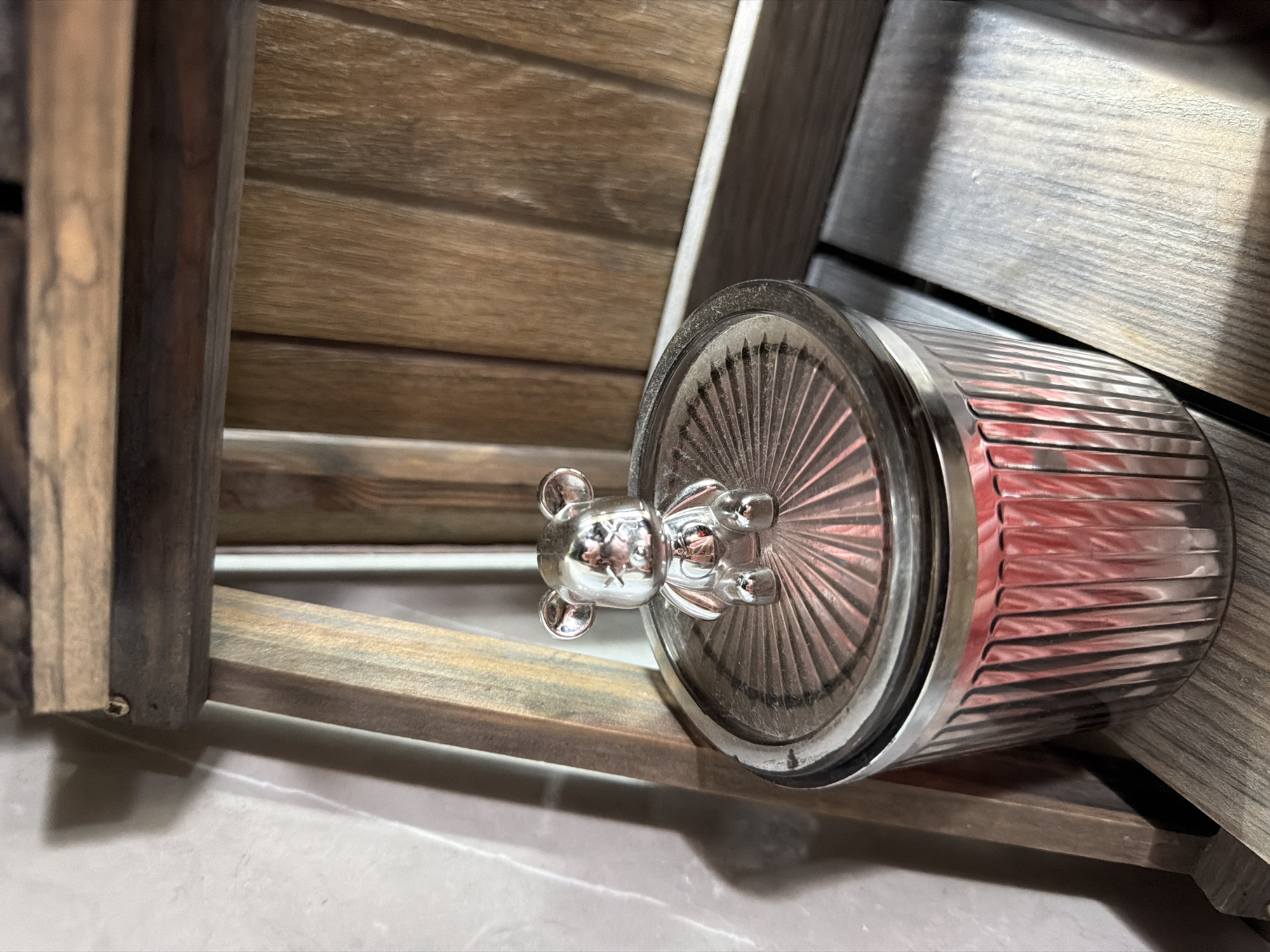 A small, shiny, silver-colored bear figurine is perched on the lid of a clear, fluted glass jar. The lid has a ribbed pattern radiating from the center. Inside the jar, pink objects are visible. The jar sits on a wooden surface, and behind it, a wooden structure with vertical slats is visible. To the left, a textured, light-colored wall with shadows cast upon it is present. The overall scene is indoors, with artificial lighting. The location is not specified in the image, nor is there any visual information that could indicate it is in Семилуки, Russia.