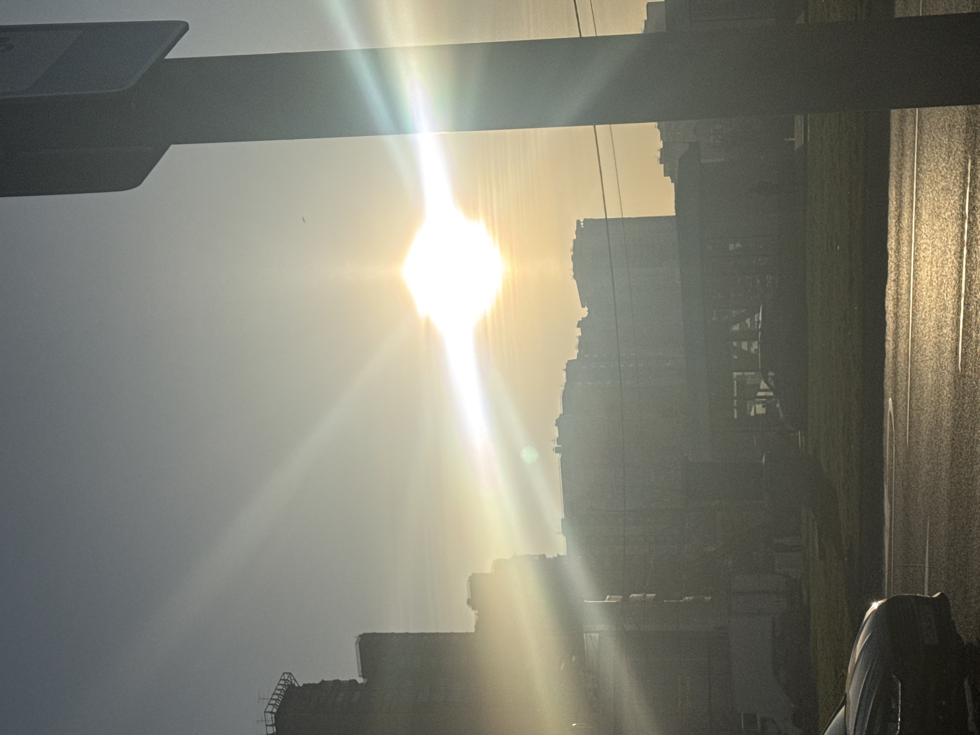 The image, taken outdoors, captures a bright, sunlit scene within a city. The sunlight, positioned centrally, radiates strongly, partially obscuring the cityscape. Buildings are silhouetted against the bright sun, their shapes and details softened by the strong light. A road or paved surface reflects the sunlight, appearing shiny. A vehicle is partially visible in the foreground. The specific location is Moscow, Russia.