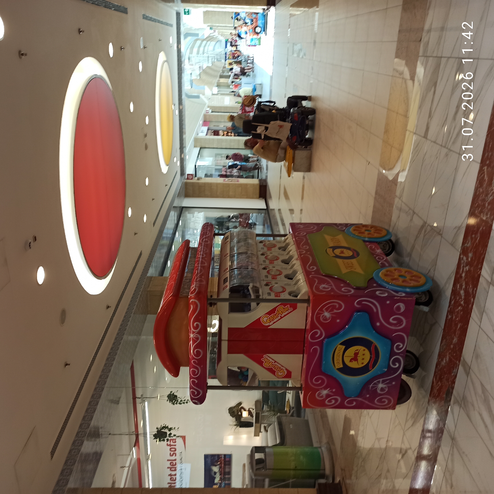 The image shows an indoor view of a shopping mall. In the foreground, a red and white candy cart with colorful decorations and wheels is prominently displayed. The cart has multiple clear dispensers filled with candy. To the left of the cart, a green and silver trash can is visible. In the background, a seating area with a yellow bench and a man in a wheelchair is visible. Further back, there are shops with large glass windows, some with merchandise displayed. People are seen walking through the mall, and in the distance, the mall extends with more shops and architectural features, including circular ceiling lights in red and yellow. The floor is tiled with a marble pattern. The overall scene depicts a typical day in a shopping mall environment. A timestamp "31.07.2026 11:42" is visible in the bottom right corner. This scene is consistent with the interior of the Centro Commerciale Bari Nord in Modugno, Italy.