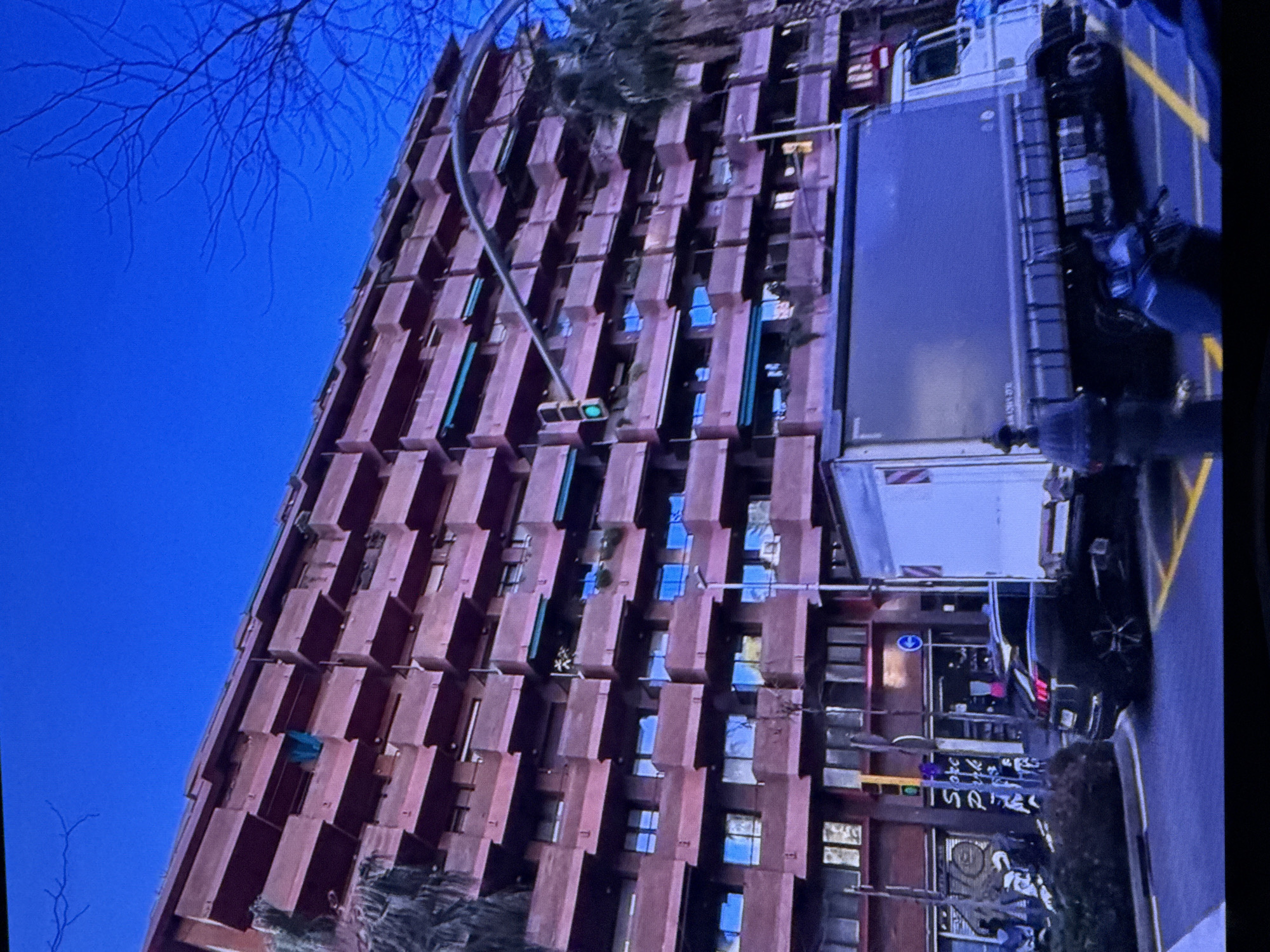 The image shows a large, multi-story residential building with a reddish-brown facade and numerous balconies. A blue sky is visible above. In the foreground, a large delivery truck is parked on a street. A dark-colored SUV is also visible, parked behind the truck. A traffic light is illuminated green. The scene appears to be an urban environment. This image is not located in Dubai, United Arab Emirates.