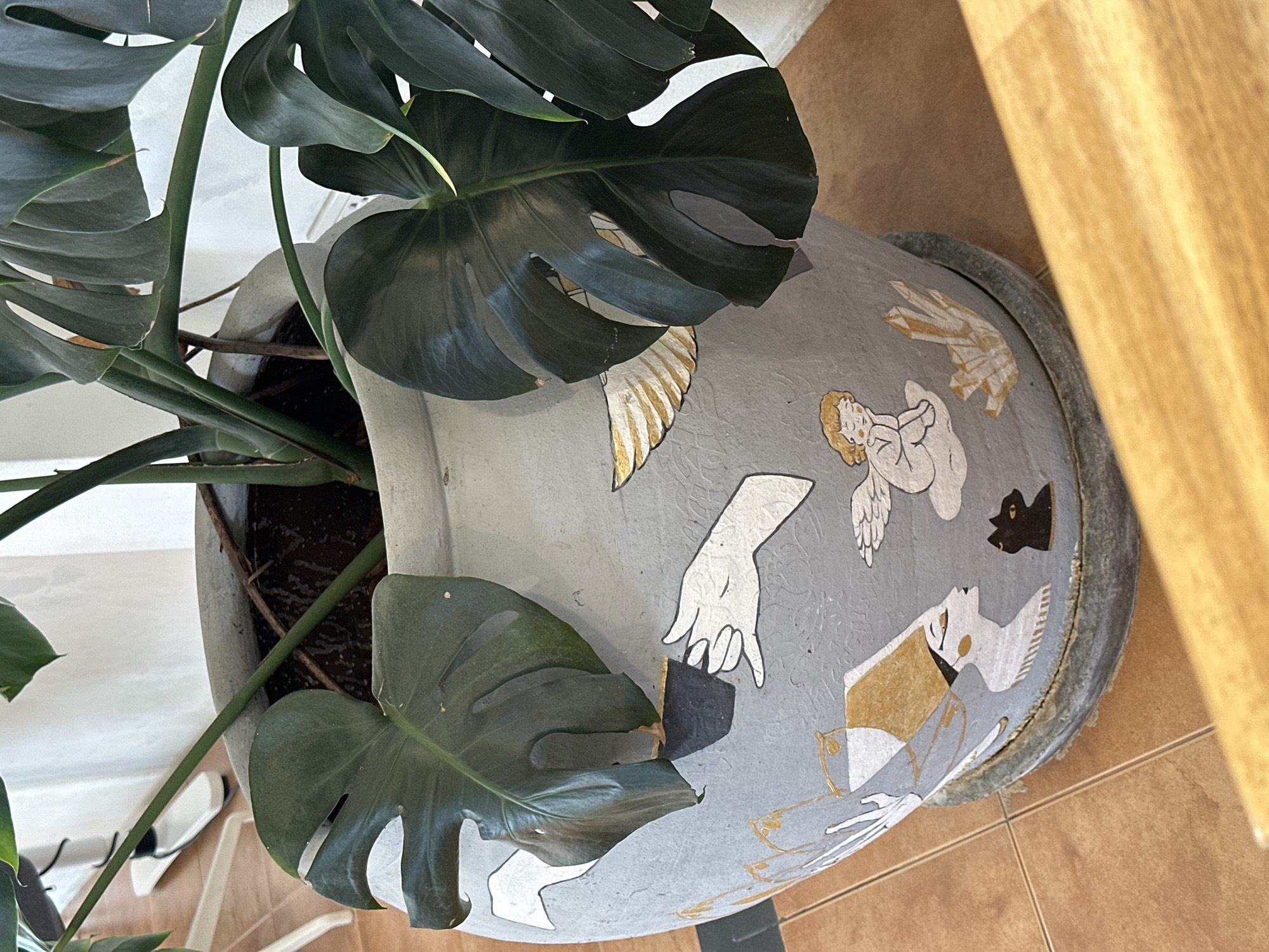 An indoor shot showcases a large, light gray plant pot with a decorative design of Egyptian figures, a black cat, an angel, and crystals. A Monstera plant with large, dark green leaves is potted within. The pot rests on a darker gray saucer, which sits on a terracotta tile floor. In the background, a white wall and a white office chair are partially visible. The scene appears to be in a residential or commercial indoor space within ตำบลเกาะพะงัน, Thailand.