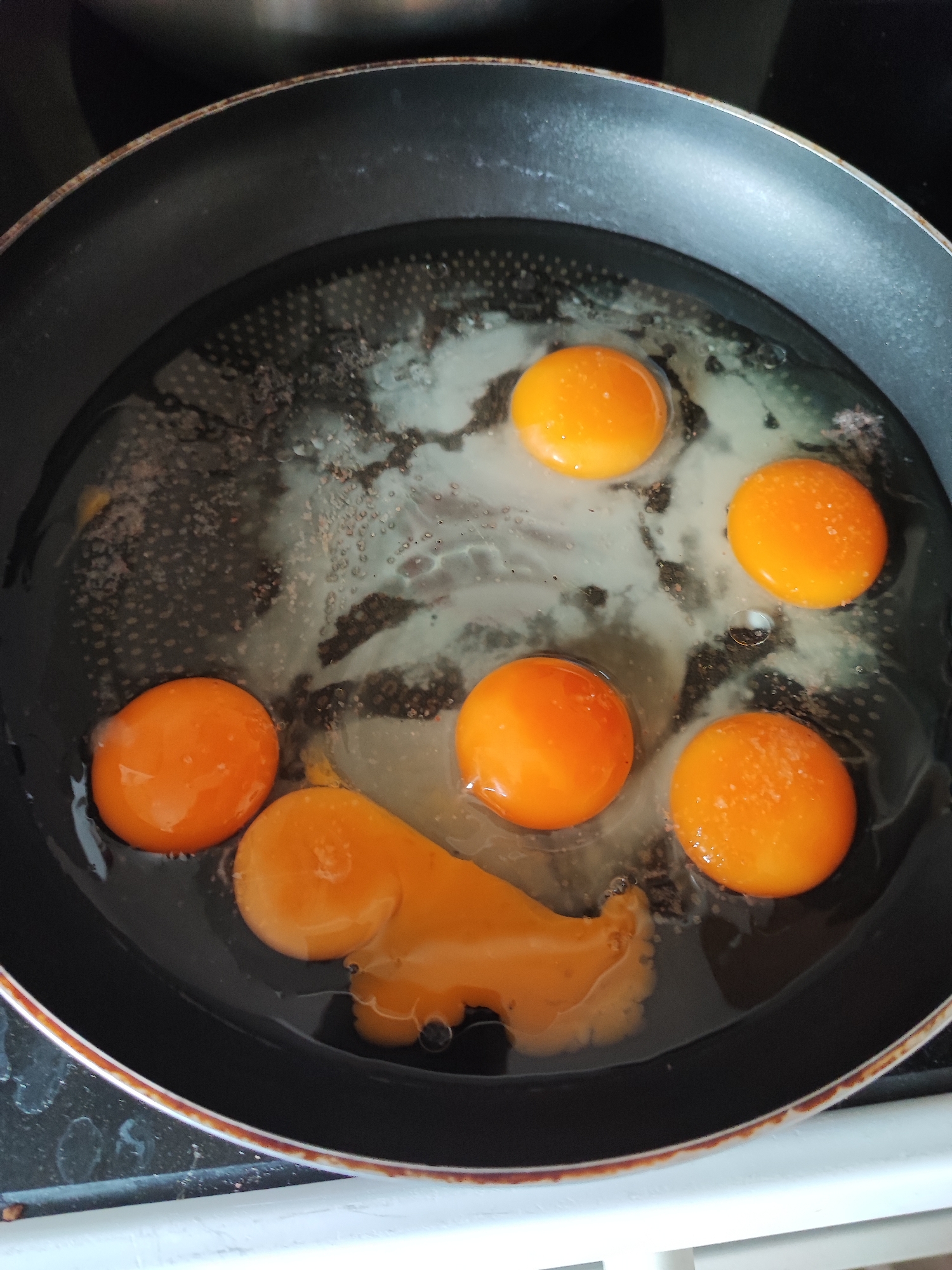 An overhead photograph depicts a black non-stick frying pan containing six raw eggs, positioned on a cooking surface within a kitchen in the city of Kadievo, Bulgaria. The pan's interior is dark with a visible dotted texture on its base, discernible through the transparent egg whites. The pan's metallic rim shows areas of brownish discoloration. Five egg yolks are intact, spherical, and exhibit a vibrant orange color. The sixth egg, located at the bottom of the pan, has a partially broken yolk that has spread into an irregular, elongated form. Small white granular speckles, consistent with seasoning, are present on several yolks and within the egg whites. The immediate background is dark, and a white horizontal surface is visible at the bottom edge of the frame.