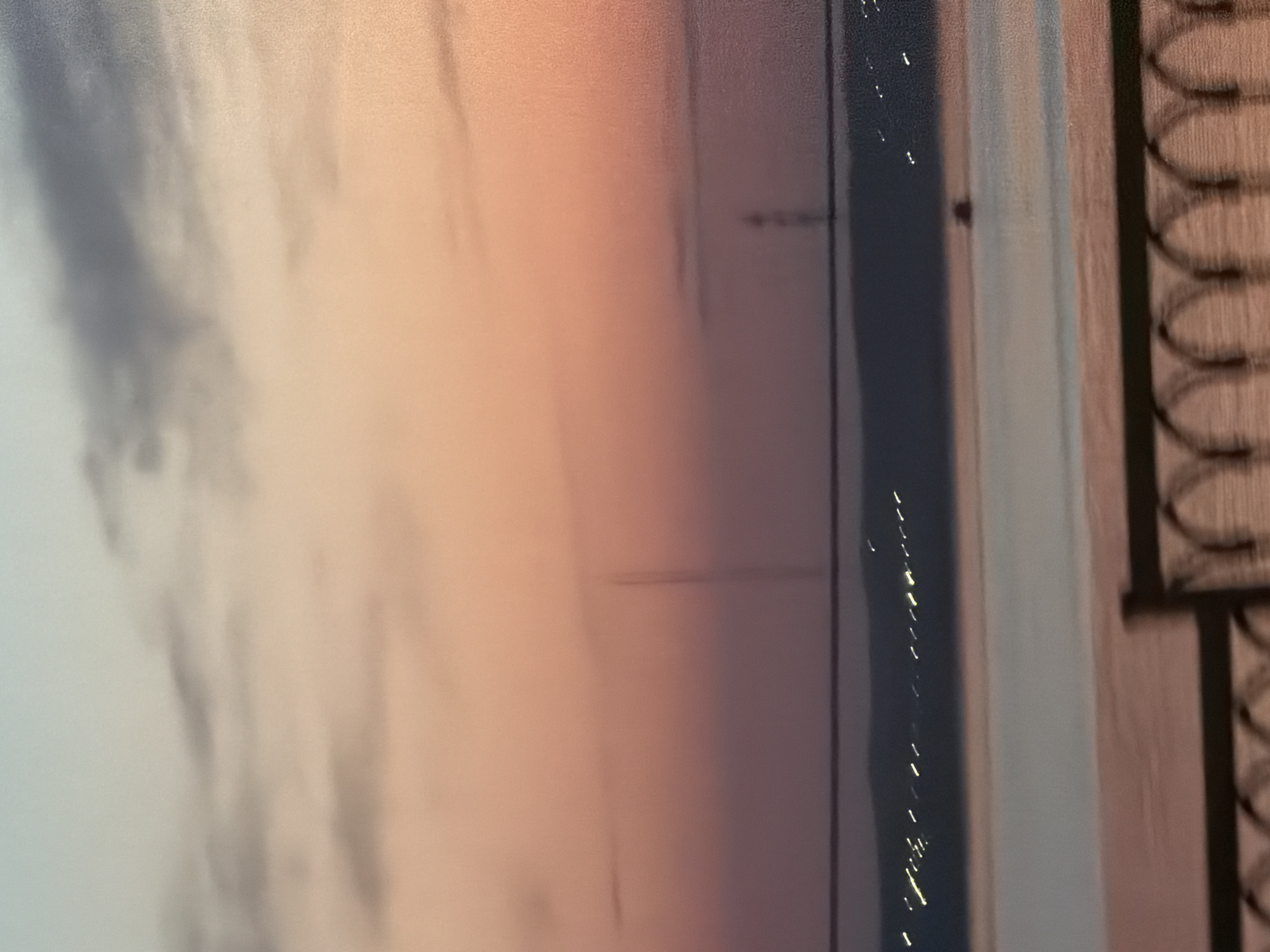 The image shows a blurry, soft-focus scene with a blend of colors ranging from light blues and grays on the left to peach and pink tones in the center. Vertical lines and dark shapes are visible on the right side, possibly suggesting architectural details like a fence or building features. The overall impression is abstract and unclear, lacking sharp focus. No specific location or people can be identified from the image.