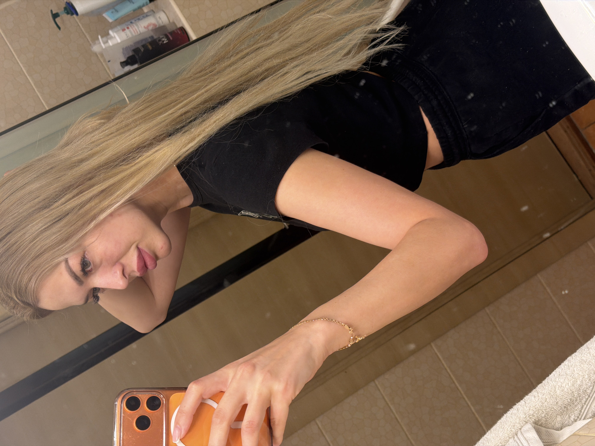 A young blonde woman takes a mirror selfie in a bathroom. She is wearing a black crop top and black sweatpants. Her left hand is holding an orange phone, and her right arm is resting on her head. The bathroom has beige tiled walls and a wooden vanity. Several bottles of toiletries are visible on a shelf behind her. The overall scene suggests a casual, personal moment captured at home.