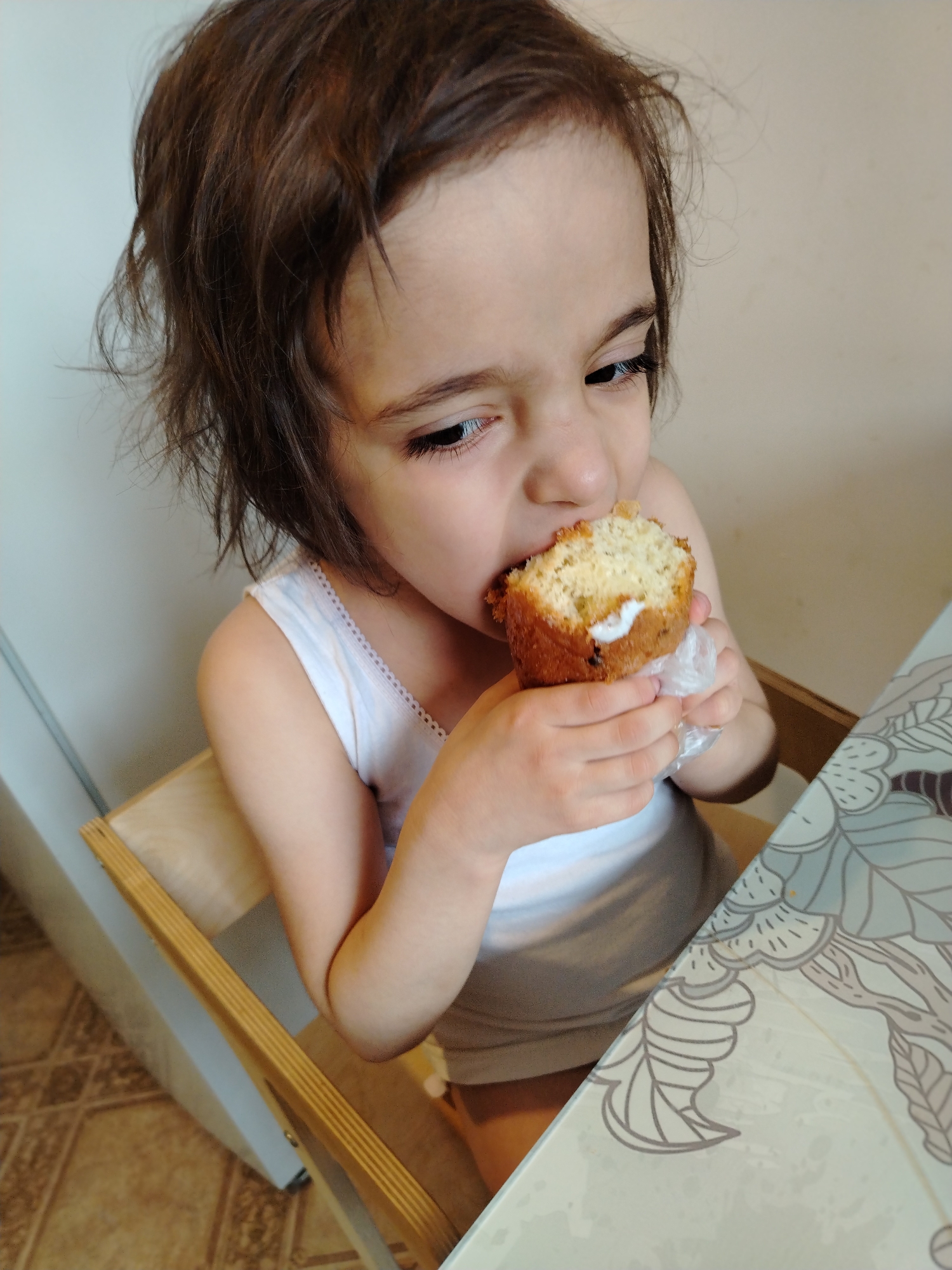 A young child with short, dark brown hair is sitting at a table and eating a muffin. The child is wearing a white tank top and is seated in a wooden high chair. The muffin appears to have a filling, possibly cream or frosting. The table has a patterned surface with floral designs in muted tones. The background includes a white wall and a glimpse of a tiled floor. The location is not specified, Noviy, Russia is not apparent from the picture.