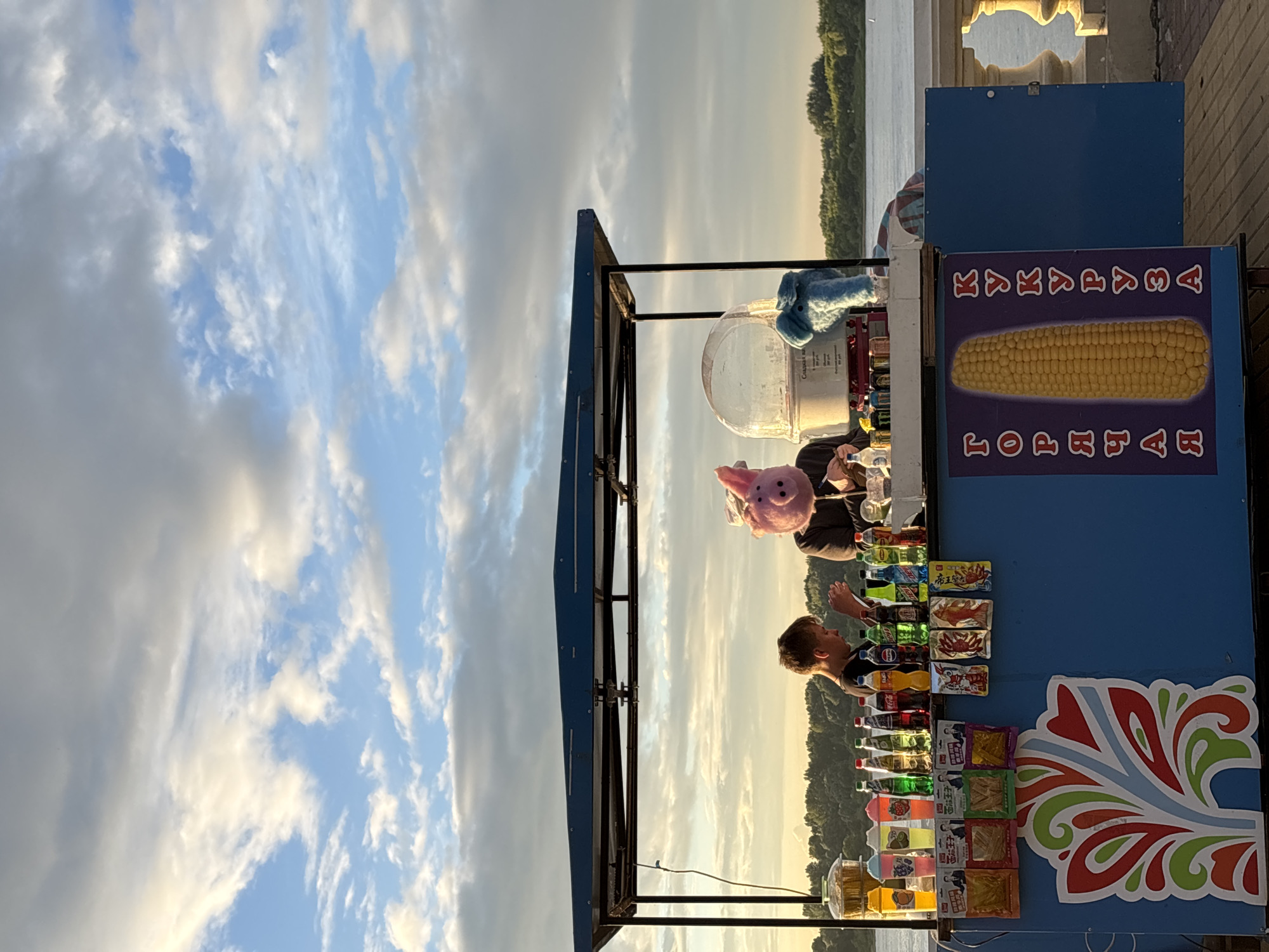 This image depicts a waterfront scene at sunset in Nizhny Novgorod, Russia. A blue cart with a dark blue awning serves as a vendor stall. Two individuals are visible behind the counter. The person on the left, a young boy, is looking down at something he is holding. The person on the right is wearing a pink pig mask and appears to be making cotton candy, with a cotton candy machine and a blue stuffed elephant also on the counter. The cart is stocked with various drinks and snacks, including brightly colored beverages in plastic cups and bottled drinks. The front of the cart features colorful signage with images of corn and text in Russian. The background shows a wide river with a tree-lined shore in the distance, and a cloudy sky illuminated by the setting sun. A stone balustrade is visible to the right of the cart. The overall scene suggests a recreational area, possibly a promenade or park along the Volga River.