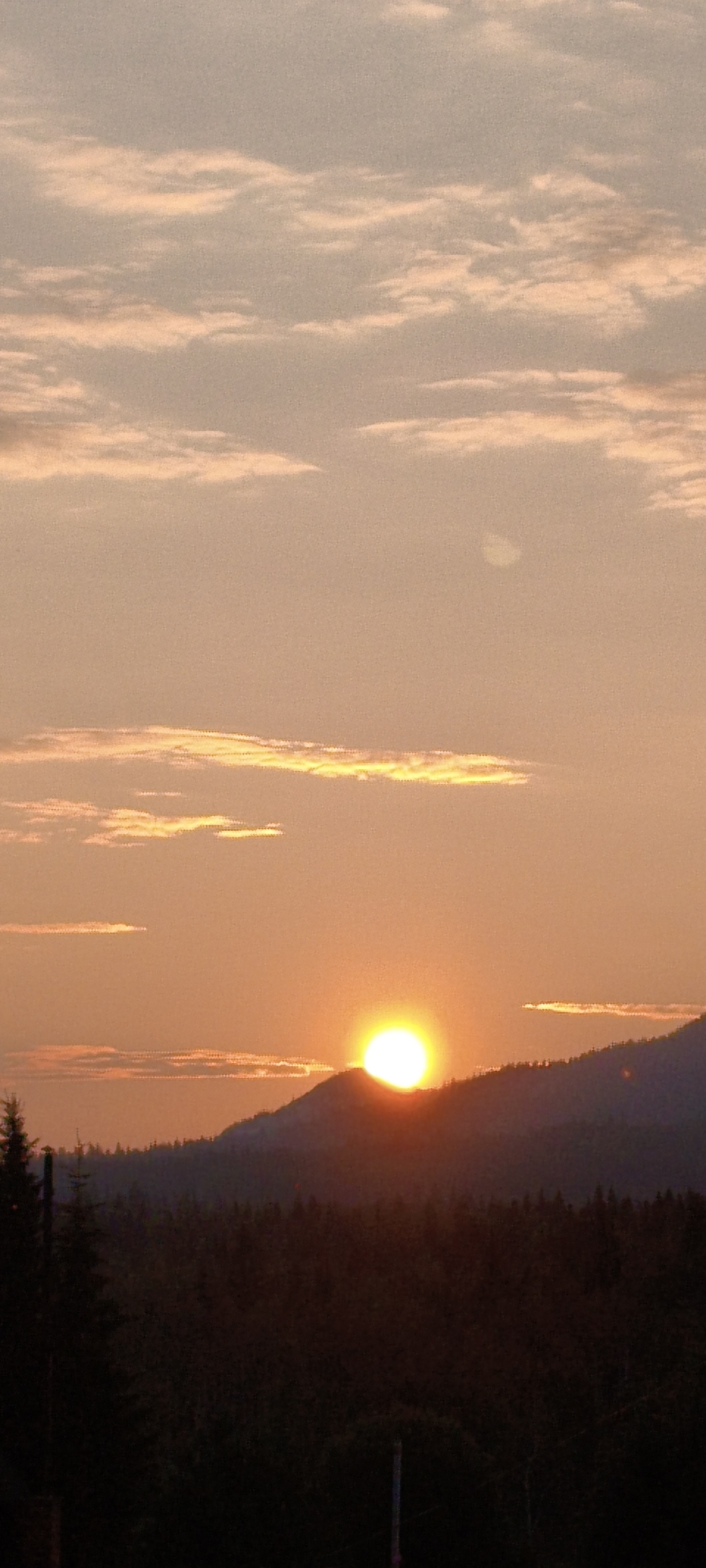 The image depicts a sunset over a mountainous and forested landscape. The sun is a bright orange orb partially obscured by the silhouette of a mountain ridge. The sky is filled with wispy clouds tinged with orange and pink hues. In the foreground, a dark, dense forest stretches across the frame. The scene is located in the vicinity of Zyuratkul', Russia, a national park known for its natural beauty and mountain ranges. There are no people or specific actions visible in the image, it is a static landscape scene.