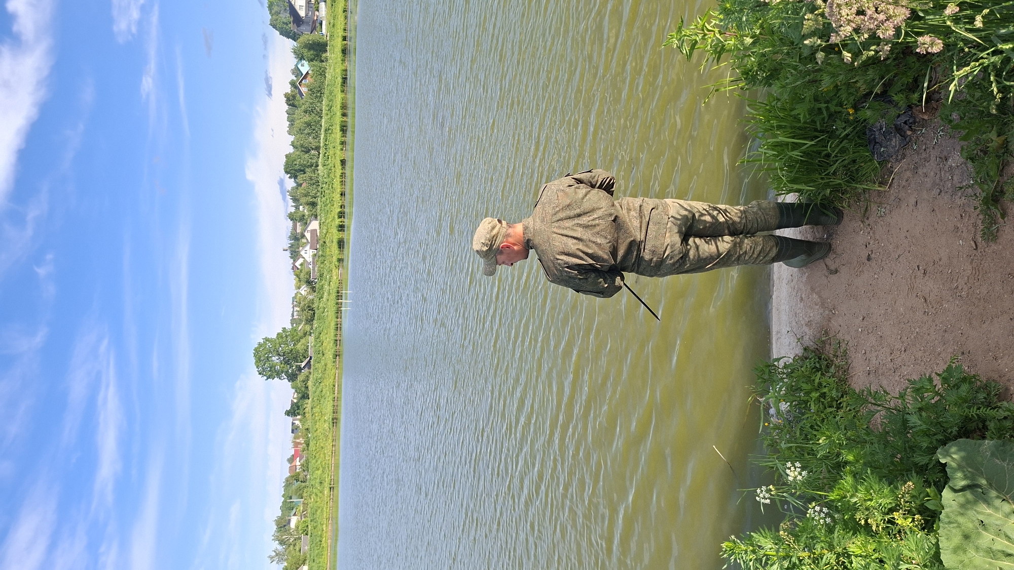A man dressed in full camouflage attire, including a jacket, trousers, and a cap, along with green boots, stands on the sandy bank of a calm body of water. He is positioned at the water's edge, looking intently downwards into the greenish-brown water. He holds a dark stick or pole, which extends from his hands towards the water surface.
The scene unfolds under a bright blue sky dotted with scattered white clouds, indicating clear, sunny weather during the daytime, likely mid-day. The body of water, possibly a lake or large pond, has a slightly rippled surface. The bank where the man stands is a mix of sand and dirt, with dense green foliage and flowering plants growing along the edge and further back. In the background, a row of green trees lines the far side of the water, behind which are visible several scattered houses and buildings, suggesting a rural village or settlement. The setting is a peaceful outdoor environment, located in the Забалоцкі сельскі Савет, Belarus. No text is visible in the image.