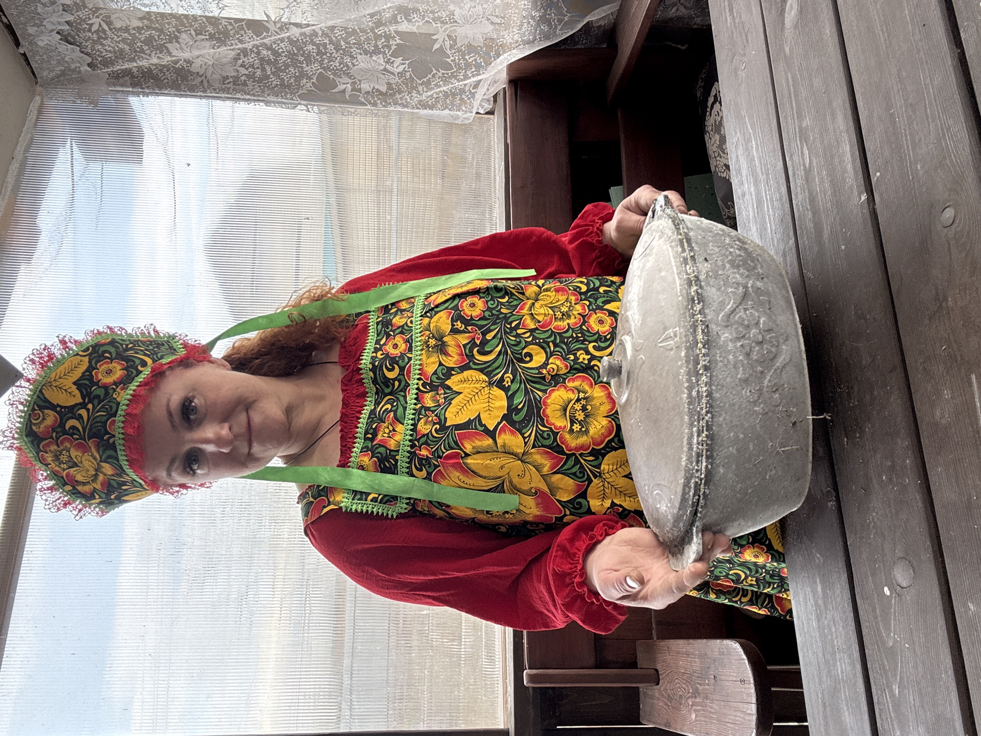 A woman wearing traditional Russian folk attire is depicted in an enclosed outdoor space, likely a veranda or greenhouse, in Chaikovsky, Russia. She has reddish-brown hair and a soft smile, looking towards the camera. Her ensemble consists of a red long-sleeved blouse with ruffled cuffs, a vibrant sarafan (apron dress) with a black background and elaborate floral patterns in yellow, orange, and green, and a matching kokoshnik (headdress) trimmed with red lace.
The woman holds a large, grey, rustic pot or dish with both hands, presenting it to the viewer. The pot appears to be made of ceramic or cast iron and features embossed floral or decorative patterns on its surface.
The background shows a transparent, ribbed panel, likely a window or wall, through which diffused daylight enters. A sheer white lace curtain with floral motifs hangs at the top of the frame. To the right, dark wooden planks suggest a table or bench. The overall scene conveys a rustic and culturally rich atmosphere. No visible text is present in the image.