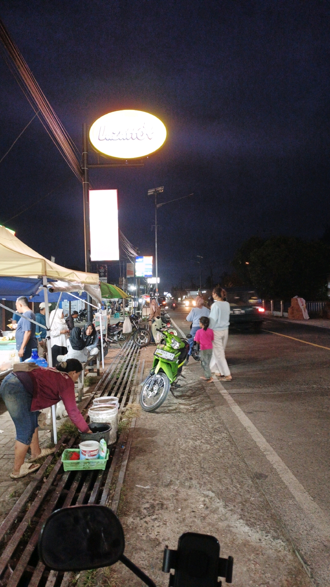 The image depicts a street scene at night in Cibenda, Indonesia, likely near a commercial area given the signage and stalls. In the foreground, a woman in a maroon top and blue shorts is bending over, appearing to wash or clean something in a green plastic container placed on a grate. Several white and black buckets are also visible next to the grate. A green motorcycle is parked on the side of the road. Further down the street, a few individuals are standing, including a woman in a light blue top and light-colored pants with a young girl in a pink top. To the left, a white tent structure with visible poles suggests a temporary stall or market. Overhead, a prominent oval-shaped illuminated sign displays the word "Lazatto's" in cursive. Other illuminated signs are visible further down the street, and traffic can be seen in the background. The overall environment appears to be an urban or semi-urban setting with ongoing commercial activity at night.