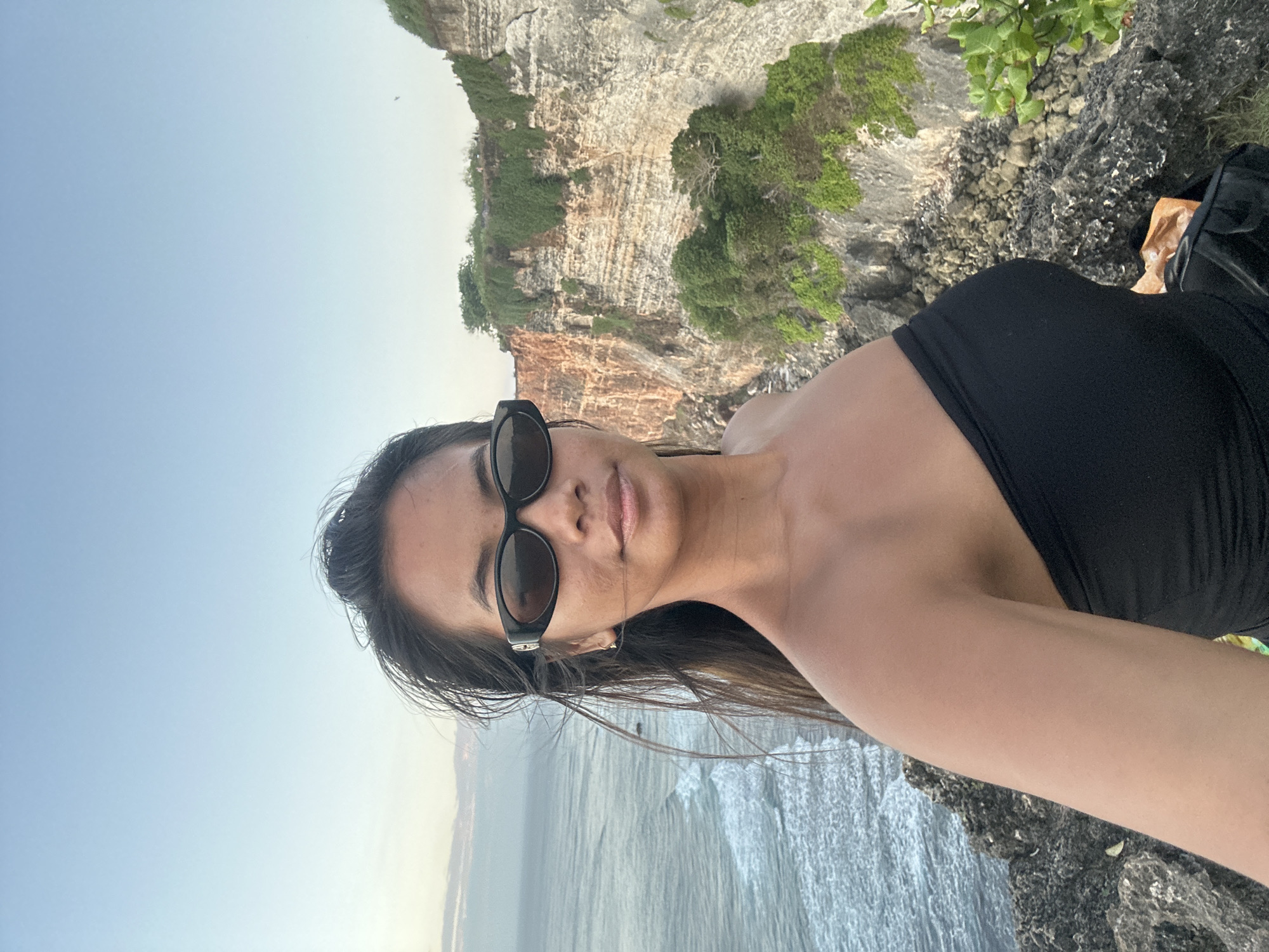 A young woman with long, dark hair pulled back and sunglasses on her face takes a selfie from a cliff overlooking the ocean. She is wearing a black strapless top. The ocean is to her left, with waves crashing against the rocks. To her right, a rocky cliff face covered in green vegetation rises up. The sky is a pale blue with some wispy clouds. The location is identified as the coast of Kuta Selatan, Indonesia.