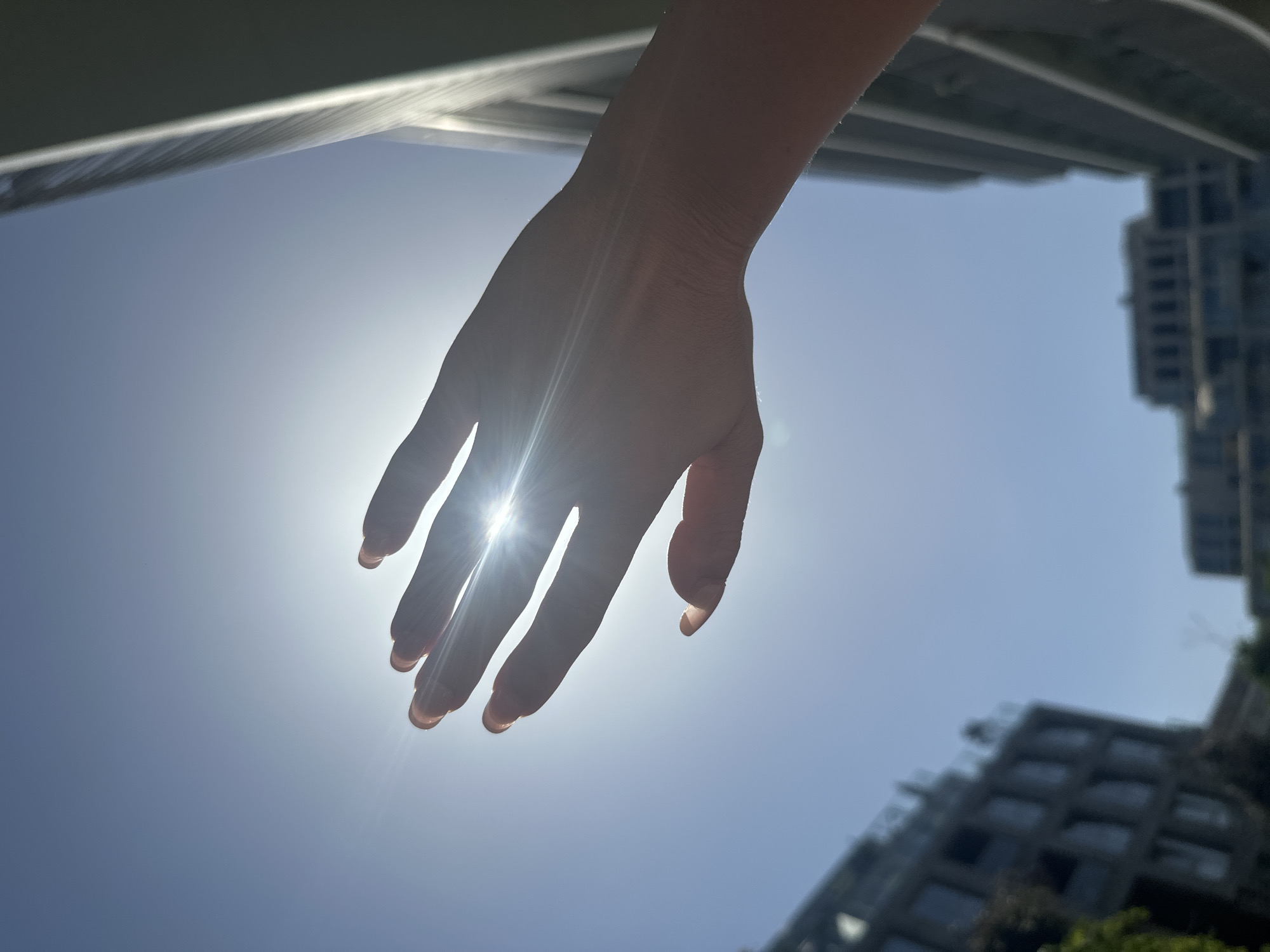 The image presents a low-angle view directed upward. A human hand, illuminated from behind by the sun, is the central focus. The sun's glare obscures the finer details of the hand but highlights its general shape and position. Behind the hand, the sky is a pale blue, indicative of daytime. At the edges of the frame are blurred outlines of buildings and architectural structures, suggesting an urban environment. The specific architectural styles are not discernible due to the blur, making precise identification of the location challenging, but they provide context to the scene.