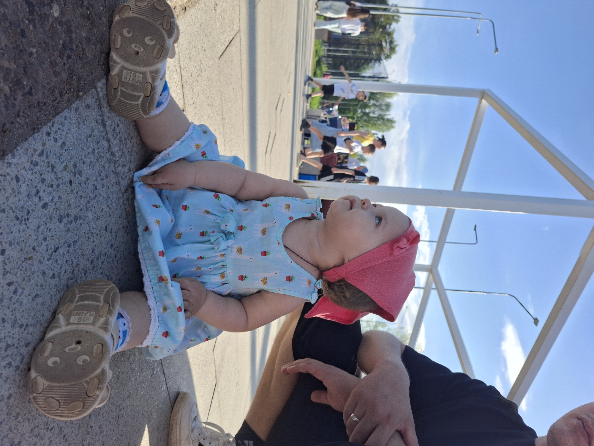 A young child, presumably a baby, sits on a concrete surface with their legs splayed. The child wears a light blue dress with a cactus pattern, white socks with blue details, and light-colored shoes with animal faces on the soles. A pink headscarf covers their head. The child is looking upwards, presumably at something out of frame.
An adult is visible behind the child, wearing black leggings and white sneakers. Only their legs and hands are partially visible.
In the background, a group of people is seen on a paved surface, near trees and street lights. Overhead, there's a white metal structure, possibly part of a public space.
The setting is outdoors under a bright, clear sky, giving the impression of a sunny day. The location is likely in Chaikovsky, Russia.