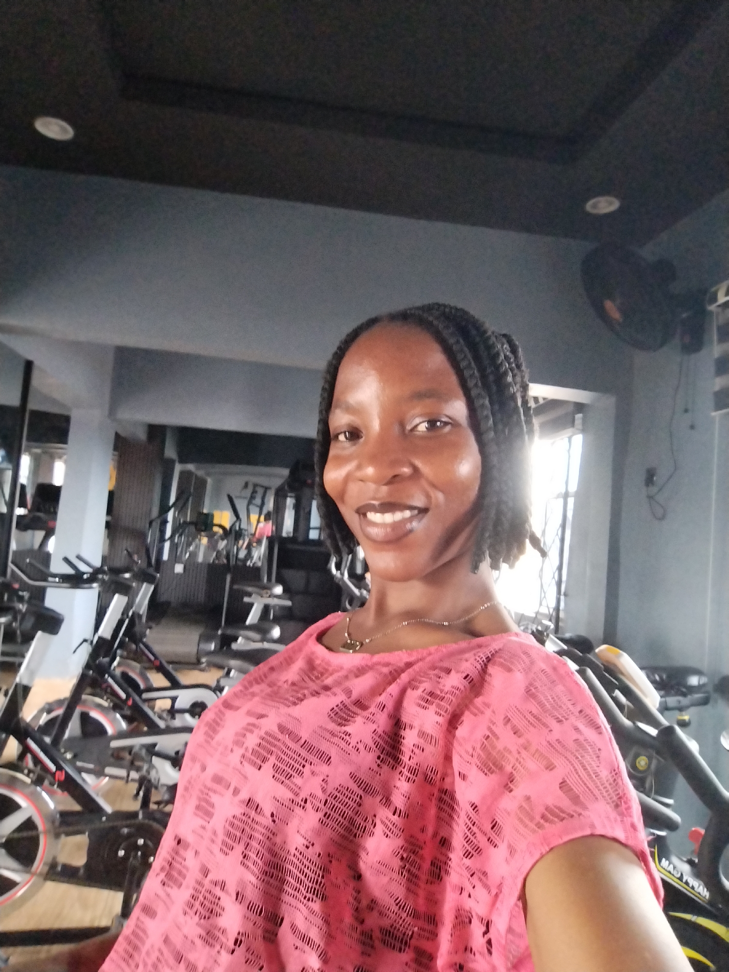 A medium shot captures a woman with braided hair smiling as she takes a selfie in a gym in Iguosa, Nigeria. She is wearing a pink, lacy, short-sleeved shirt and a delicate silver necklace. The background is filled with various exercise machines, including several stationary bikes. The lighting suggests it is daytime, with natural light coming from a window or door in the background. The gym appears to be well-equipped.