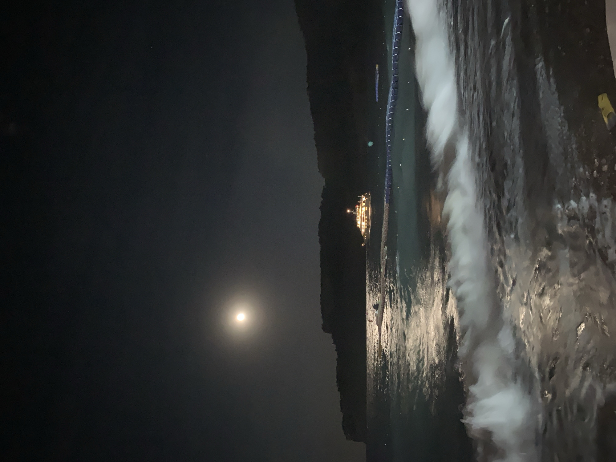 An outdoor nighttime scene captures a moonlit body of water, likely the Mediterranean Sea, with a dark, tree-covered hillside in the background. A full moon, illuminated by a soft glow, hangs in the dark sky, reflecting its light onto the water's surface. Gentle waves crash onto the shore, creating a foamy white texture in the foreground.
In the middle ground, a large, lit-up boat, possibly a yacht, is anchored offshore. Adjacent to the boat, a floating barrier with blue lights stretches across the water, indicating a designated swimming or activity area. A small inflatable boat with a red marker is tied to the floating barrier. The lights on the barrier and boat create a festive atmosphere against the dark night. The scene is likely located in the coastal city of Kemer, Turkey, known for its beautiful coastline and nighttime activities.