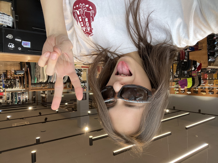 A young female, with long brown hair, poses for a self-portrait within a retail store. She wears dark, sport-style sunglasses and a white t-shirt featuring a red graphic resembling a stylized building or tower. Her left hand is raised, displaying a peace sign, and her tongue is extended. Her right eye, partially visible behind the sunglasses, appears to be squinted or winking.
The background reveals a well-lit retail environment. The ceiling is light-colored, featuring multiple black linear LED lighting fixtures and black track lights with individual spotlights. Merchandise is displayed throughout the store: footwear (athletic shoes/sneakers) on wooden shelving units along the left wall, various hats (including neon yellow and purple beanies) on display racks, and clothing on hanging racks further back. Vertically oriented snowboards or longboards are visible on a display rack on the far right wall. A black display counter with various stickers, including a recognizable Vans logo, is present in the foreground on the right.
The interior architecture and merchandise presentation are consistent with a "Traektoria" (Траектория) extreme sports and streetwear store, identifying the location as likely a "Traektoria" branch in Krasnoyarsk, Russia.