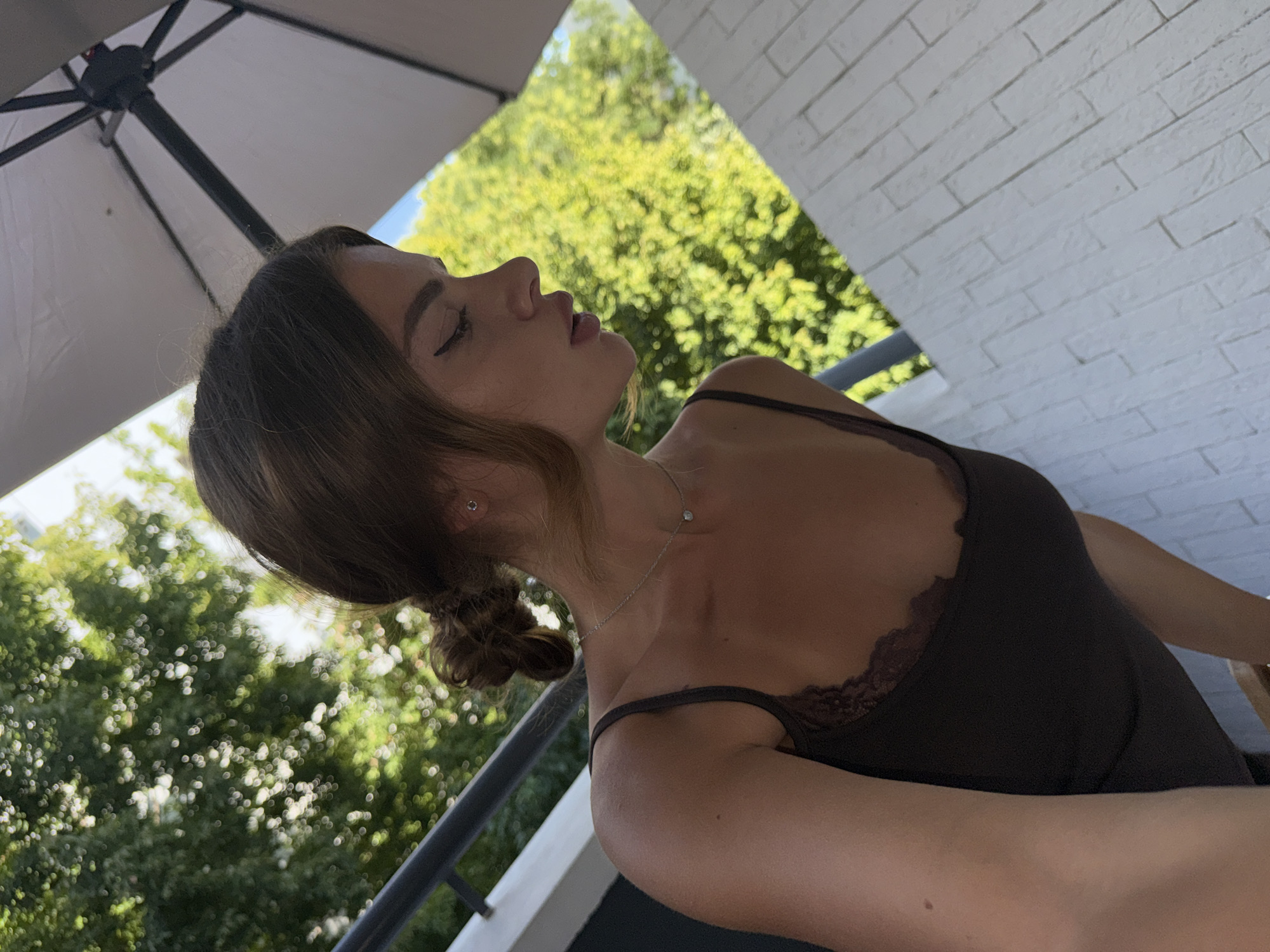 The image features a young woman posing outdoors. She is facing away from the camera, with her head tilted back and eyes closed, suggesting a moment of relaxation or contemplation. Her hair is styled in two buns. She is wearing a dark camisole with lace trim and a delicate silver necklace with a small pendant. The background consists of lush green trees, indicating a natural setting, possibly a garden or park, bathed in natural light, suggesting it is daytime. To the right, a white brick wall is visible, and an outdoor umbrella is partially in the frame on the left. The scene appears to be set in a warm climate, possibly during summer, given the foliage and lighting. The location is identified as Plovdiv, Bulgaria. There is no visible text in the image.