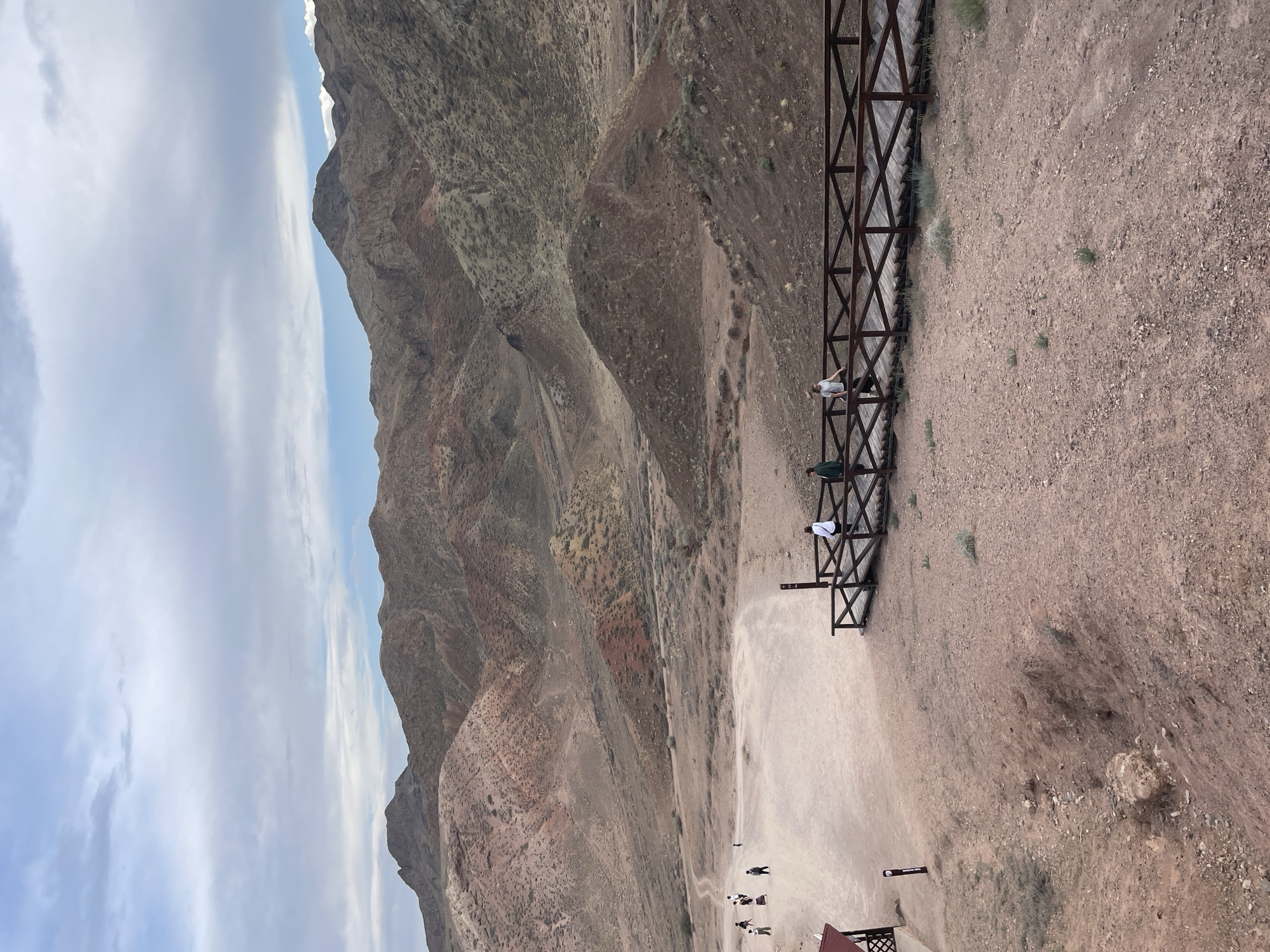 This image captures a scenic view of the Charyn National Park near the city of Tamerlik, Kazakhstan. In the foreground, a wooden boardwalk with railing allows visitors to traverse the sandy, rocky terrain. Three individuals are visible on the boardwalk, two appearing to walk away from the camera and one facing the landscape. Further in the midground, a group of people are dispersed across a wide, sandy path, some walking, others standing. A small, red-roofed structure is present on the left side of the image, with a tall, dark signpost nearby. The midground transitions into a vast, arid landscape characterized by rolling hills and mountains. These geological formations exhibit a striking palette of earthy tones, ranging from reddish-browns and ochres to muted grays and greens. Vegetation is sparse, consisting mainly of low-lying shrubs and grasses clinging to the slopes. The background is dominated by a range of imposing mountains, their rugged contours softened by distance and atmospheric haze. The sky above is partly cloudy, with a mix of light gray and white clouds against a pale blue backdrop. The overall impression is one of a remote, natural environment, likely a tourist destination known for its unique geological features.