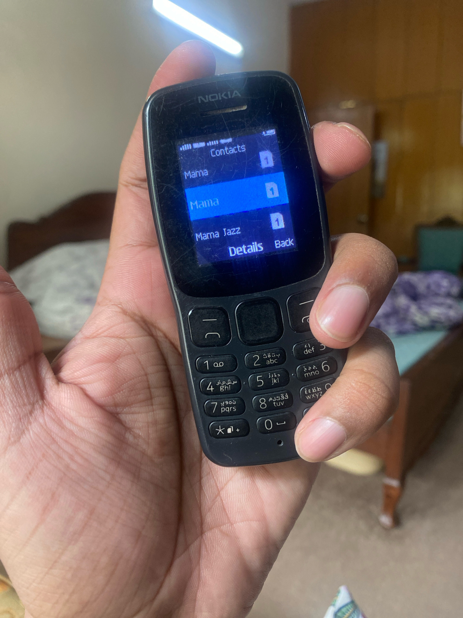 A Nokia 105 mobile phone is held in a person's left hand. The phone's screen displays a contacts list with entries for "Mama", "Mama", and "Mama Jazz". The surrounding environment is blurry, but appears to be an indoor setting with a bed visible in the background, suggesting a domestic environment. The image does not provide any information about the location being Keer, Pakistan.