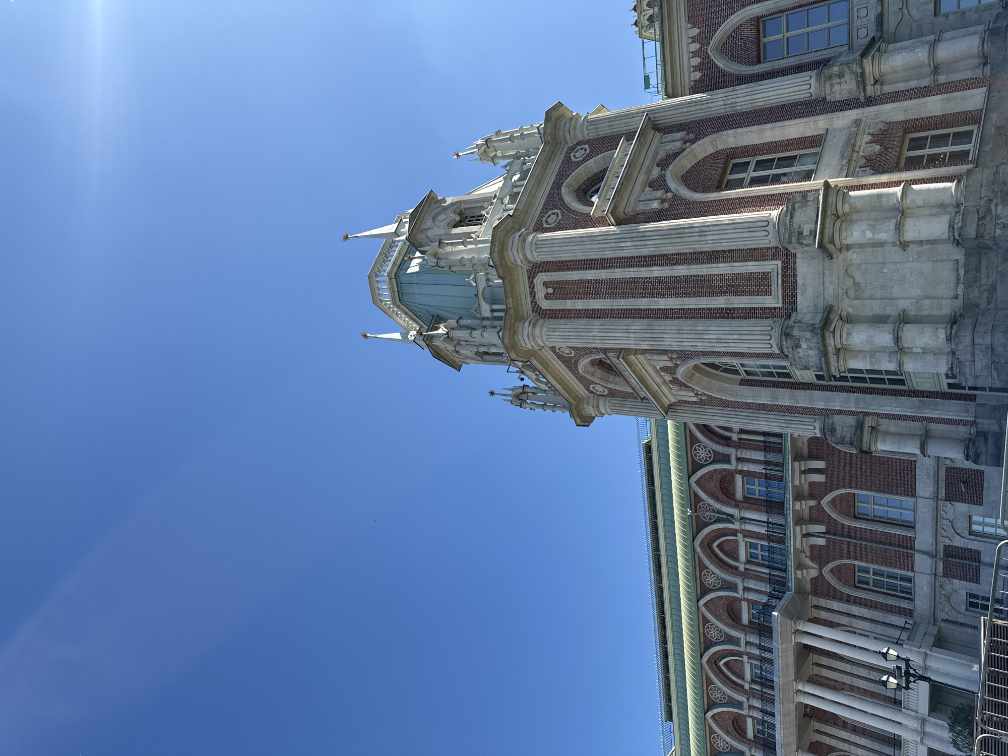 The image shows the Tsaritsyno Palace in Moscow, Russia, on a clear, sunny day. The palace is a complex of buildings in a Neo-Gothic architectural style, characterized by red brick facades, white stone detailing, and ornate spires. The photograph is taken from a low angle, emphasizing the height and grandeur of the central tower. The sky is a brilliant, cloudless blue, with a slight lens flare visible in the upper right corner, indicating the sun's position. The palace's intricate details, including arched windows, decorative carvings, and ribbed columns, are clearly visible. The surrounding foliage or park area is not prominently featured, with the focus solely on the majestic architecture against the vast expanse of the sky.