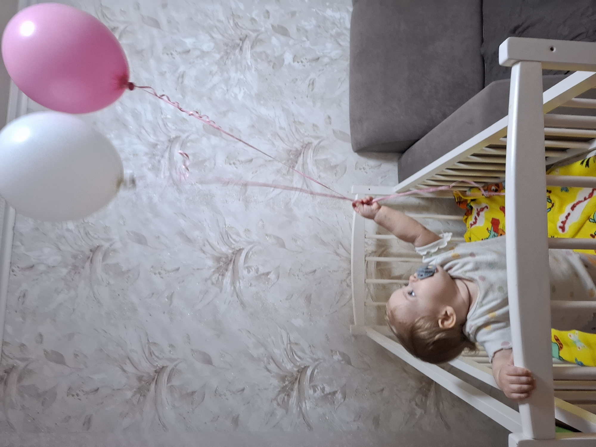 An indoor, eye-level shot captures a baby peering over the side of a white wooden crib, reaching towards two floating balloons. The image is taken at a ninety-degree angle, making the baby appear to be lying on their side as they lean out. The baby has light hair, is wearing a light-colored onesie, and has a blue pacifier in their mouth. Their right hand is extended, gripping the thin, curly pink ribbons attached to the balloons. A yellow blanket or clothing with a cartoon pattern is visible inside the crib.
Two balloons are prominent in the upper left portion of the frame: one bright pink and one white, both held by the same pink ribbons. They float against a wall covered in light-toned wallpaper, featuring a subtle, abstract floral or leafy pattern in grays and whites. In the background, behind the crib, a part of a dark gray upholstered sofa or headboard is visible. The scene appears to be set in a home, possibly a nursery or living room, located in Chaikovsky, Russia, based on the provided context. The baby looks curious and engaged with the balloons, suggesting a playful moment. There is no visible text in the image.