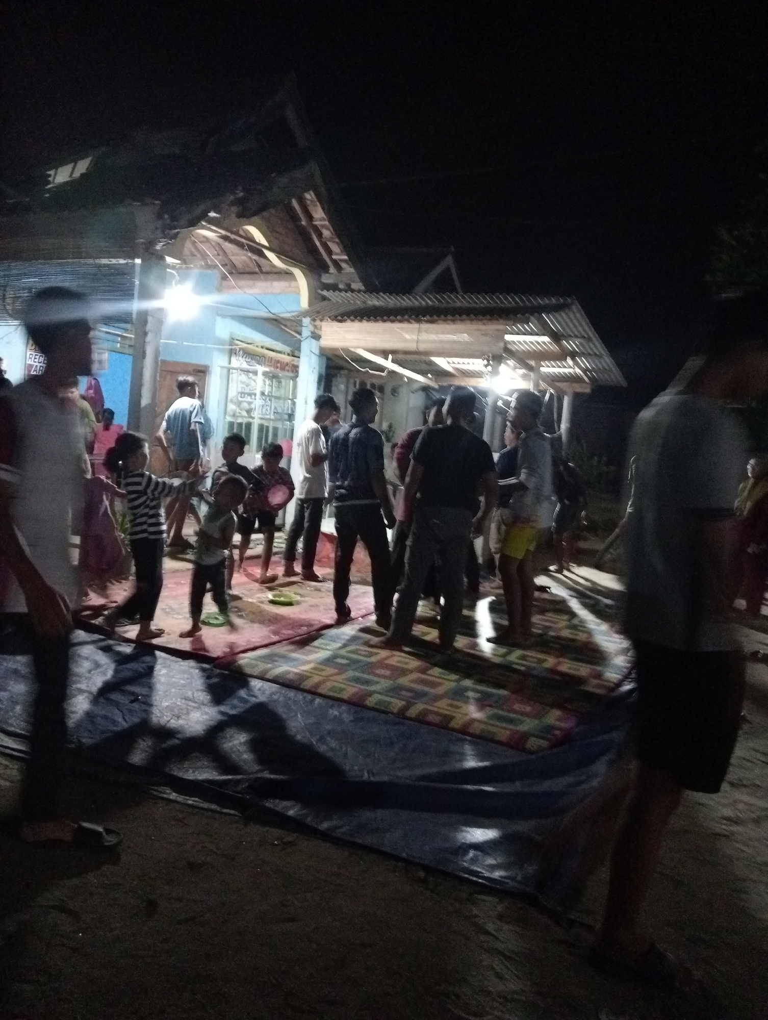 The image depicts an outdoor gathering at night in Rasuan Baru, Indonesia. Several individuals, including adults and children, are present. They are standing and interacting on a dirt ground covered with a large blue tarp and a patterned rug. Some people are holding objects, possibly related to food or festivities. The background shows a building with a corrugated metal roof and a blue wall, illuminated by artificial lights. Shadows are cast across the ground, indicating the presence of strong light sources. The atmosphere appears to be informal and communal.