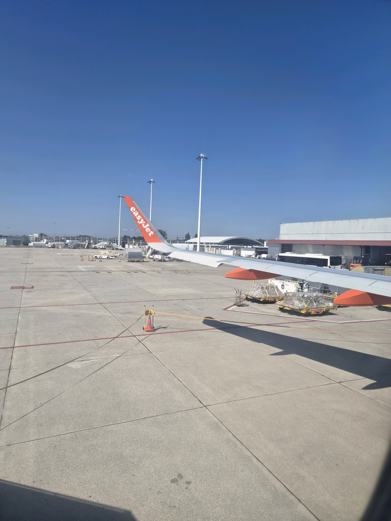 The image depicts an exterior view of an airport tarmac under a clear blue sky in Vila Nova da Telha, Portugal. In the foreground, the wing of an EasyJet airplane, with its distinctive orange logo and text, extends across the frame. The tarmac is made of concrete and marked with various lines, including red and yellow boundaries. Several airport vehicles and equipment are visible, including baggage carts loaded with cargo, some covered with netting. In the background, airport buildings and structures, including a large hangar-like building and what appears to be a bus, are present. Tall light poles are also dotted across the tarmac. The scene appears to be a typical airport environment with an airplane preparing for or having just completed a flight.