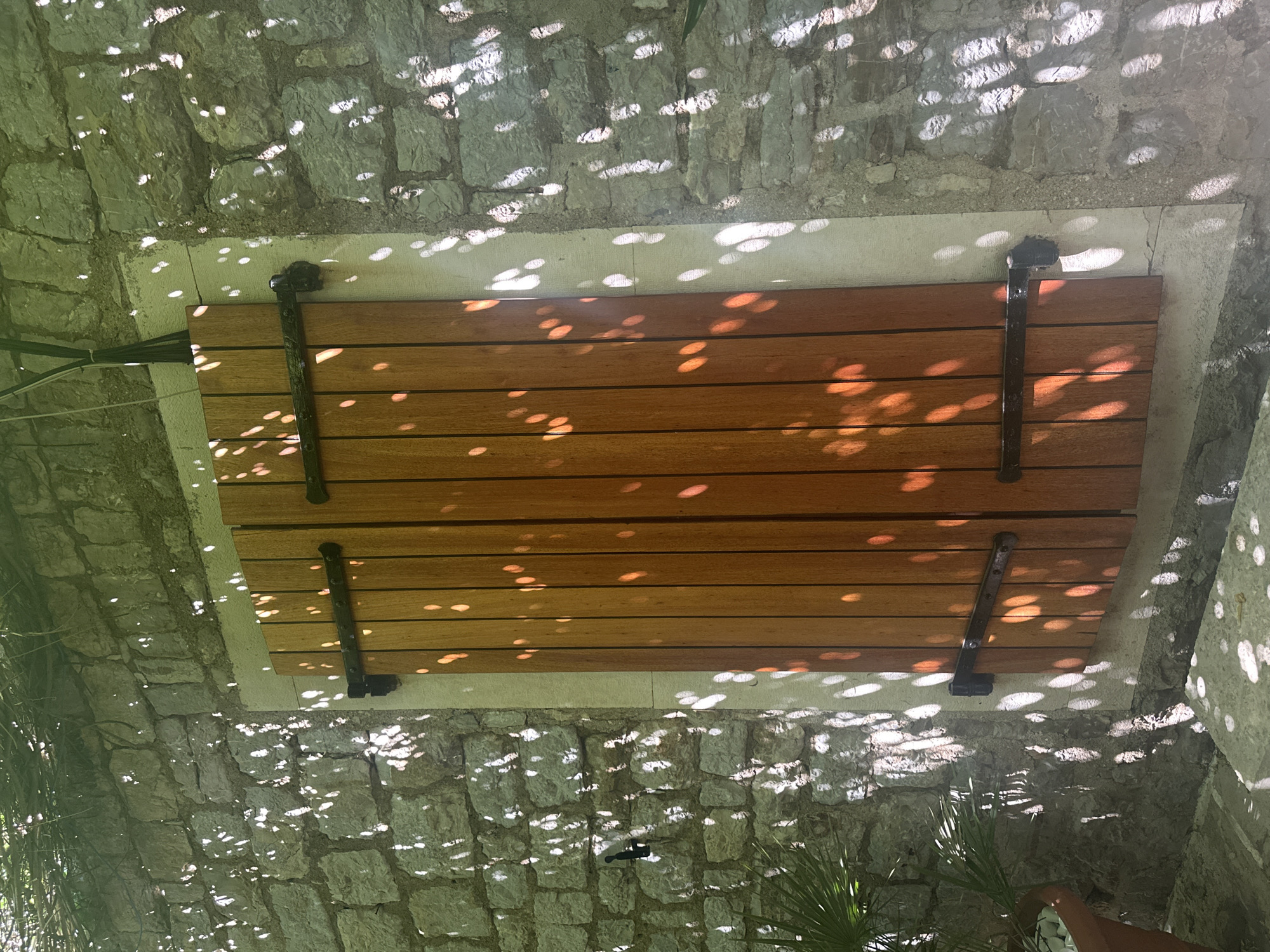 The image shows a wooden hatch or cover set into a stone wall structure. The hatch is made of horizontally aligned wooden planks with black metal hinges. The stone wall is constructed of irregular, light-colored stones, and the area is dappled with light, suggesting it is outdoors beneath foliage. The context of the city of Budva, Montenegro, is not directly visible in the image itself.