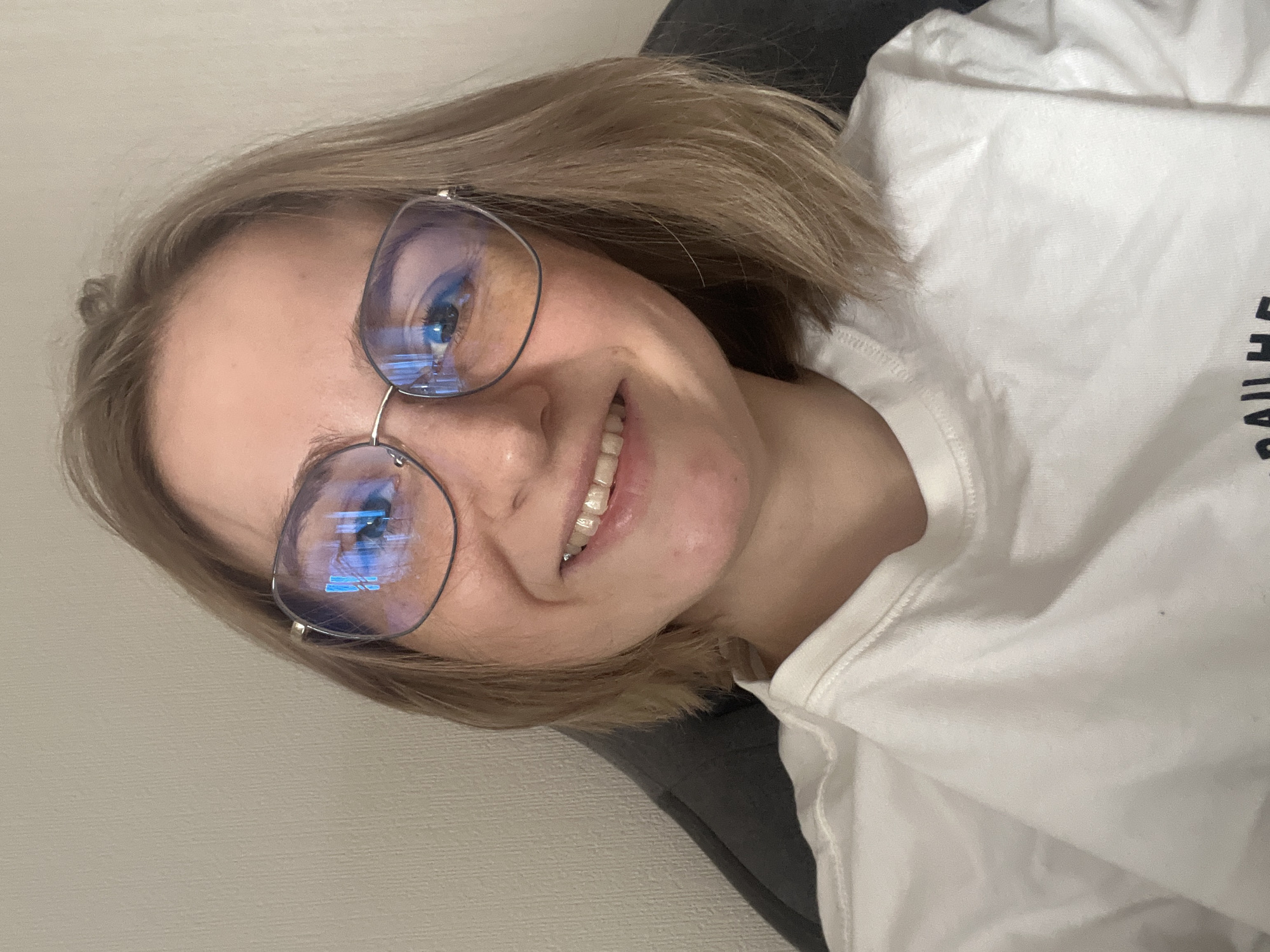 A medium close-up depicts a smiling Caucasian woman with short, light brown hair wearing silver-rimmed glasses. The glasses reflect a blue, light source. She is wearing a white t-shirt and appears to be reclining against a dark gray object or surface. The background is a light beige wall. There is no evidence in the image that indicates the location is Novosibirsk, Russia.