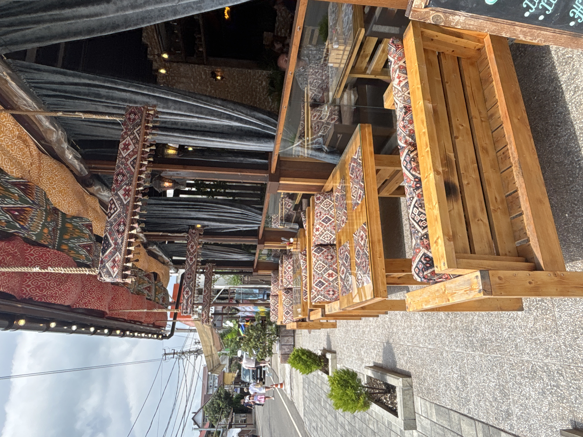 This image depicts an outdoor seating area of a cafe or restaurant, likely in Sochi, Russia, during daylight hours with a cloudy sky. The scene features several wooden tables and benches with cushions adorned with a vibrant, geometric pattern. The seating is arranged along a tiled walkway, with planters containing small, manicured evergreen bushes.
In the background, the street is visible with a few cars and pedestrians. The architecture suggests a commercial area, possibly a resort town. The cafe area itself is covered by a dark fabric awning, creating shade and a cozy ambiance. Decorative elements like tassels and patterned fabrics hang from the structure, adding to the aesthetic.
The overall impression is one of a relaxed, open-air dining or lounging space, designed to be inviting and visually appealing. The sunlight creates shadows on the ground, indicating it is likely daytime, though the cloudy sky might suggest overcast conditions. No specific emotions are conveyed by any visible subjects, as the focus is on the setting and its design. There is a chalkboard sign visible at the top right, but the text on it is illegible.