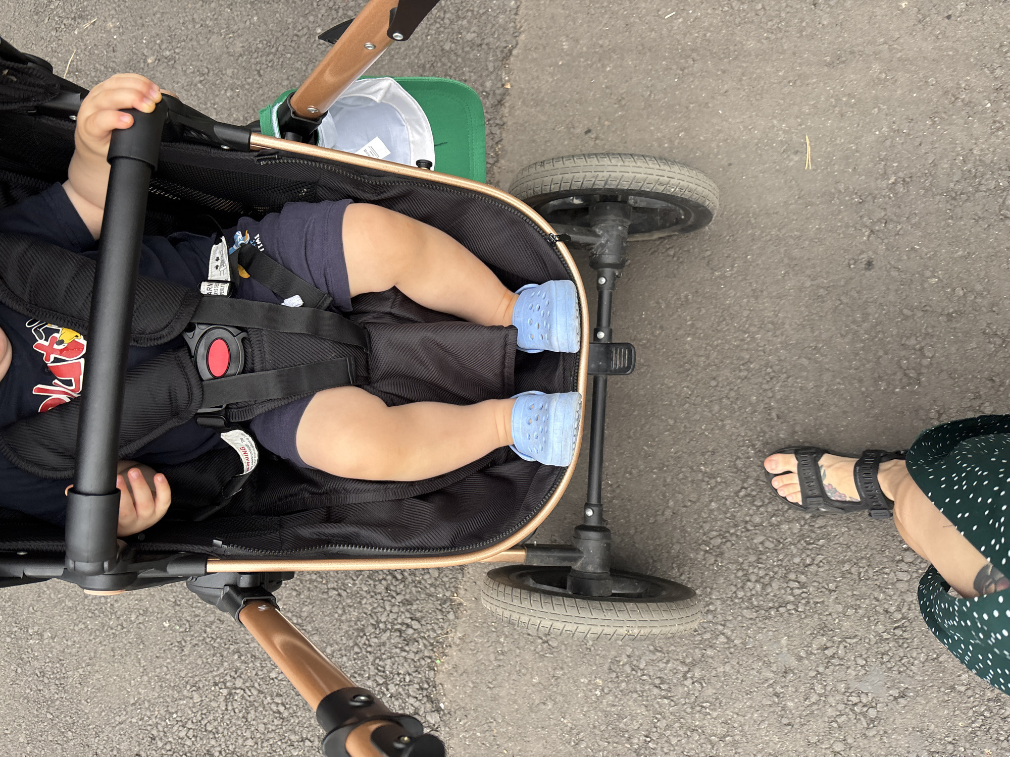 A high-angle, medium shot shows a child sitting in a stroller, with a person's legs and feet in the foreground, on a paved surface, likely in the city of Naberezhnye Chelny, Russia. The child is wearing a dark blue t-shirt with a red and yellow graphic, dark blue shorts, and light blue clogs. The child's hands are visible resting on the stroller's safety bar. The stroller has a black fabric interior, a black safety harness with a red buckle, and a bronze-colored frame and handle. A white and green baseball cap is tucked into the stroller behind the child. The person in the foreground is wearing a dark green dress with white polka dots and black sandals. The paved surface appears to be asphalt, with visible gravel and some debris. The lighting suggests it is daytime.