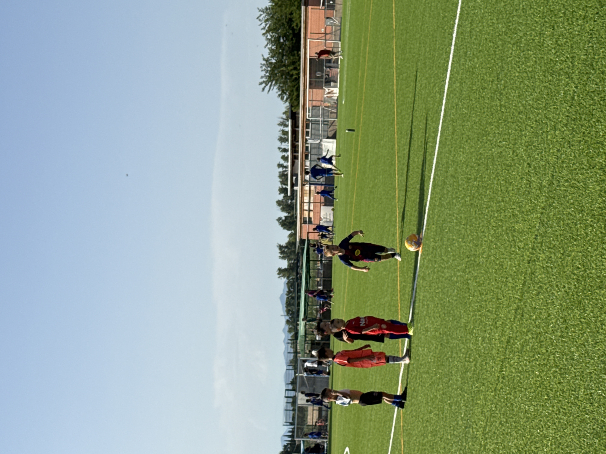 The image depicts a children's soccer practice on an artificial turf field with a light blue sky and scattered clouds. Several young boys in soccer uniforms are on the field. In the foreground, on the left, three boys stand in a line, one in a white and black striped jersey, another in an orange jersey, and a third in a red jersey. To their right, a boy in a dark blue and red jersey is running. A soccer ball is visible near the boy in the dark blue and red jersey. In the background, more children and adults are present on the field, some engaged in practice and others observing. A building with a red brick facade and white trim is situated behind the field, with trees and vegetation surrounding it. The field has white and yellow markings. Based on the context provided, this scene is located in Vidreres, Spain.