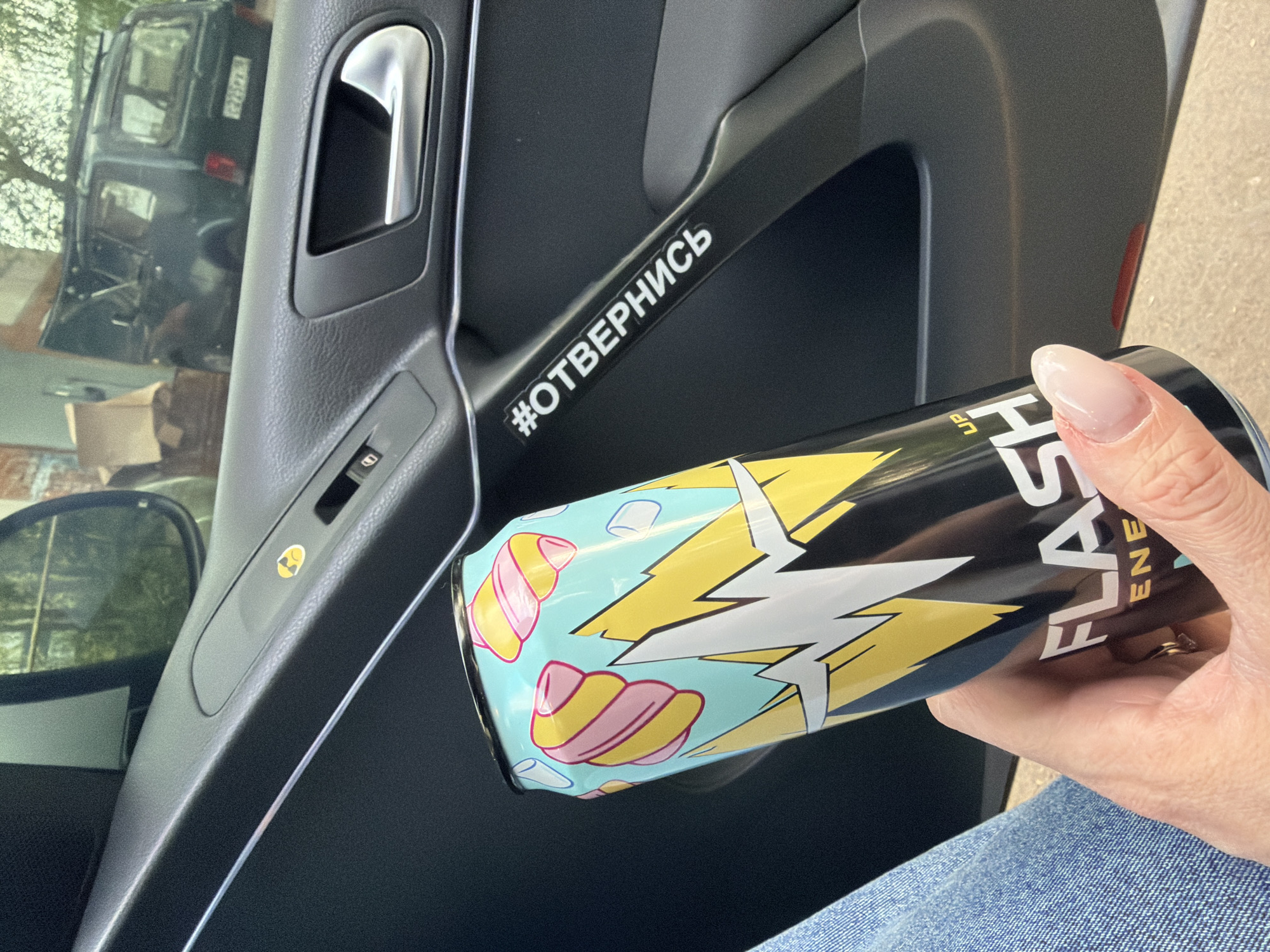 A can of Flash energy drink is held in a person's hand inside a car. The car's door panel is visible, with a sticker that reads "#отвернись". The city and specific location are unknown.