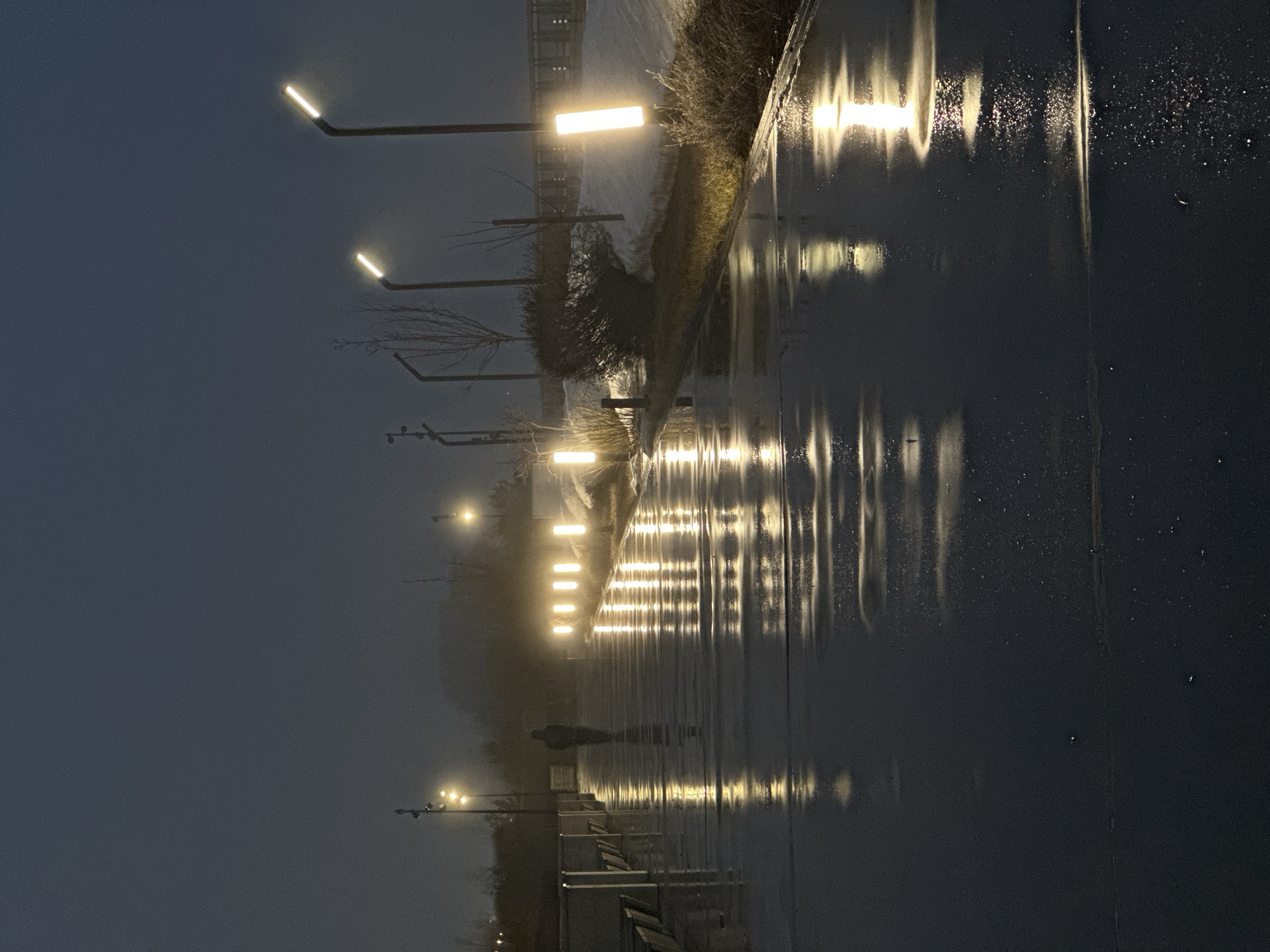 This image depicts a wet, outdoor scene at dusk in Kazan, Russia. The location is a promenade or walkway illuminated by a series of modern, rectangular streetlights. The ground is reflective, suggesting recent rain or a wet surface. A lone person walks along the promenade. Vegetation borders the walkway. The sky is overcast and dark.