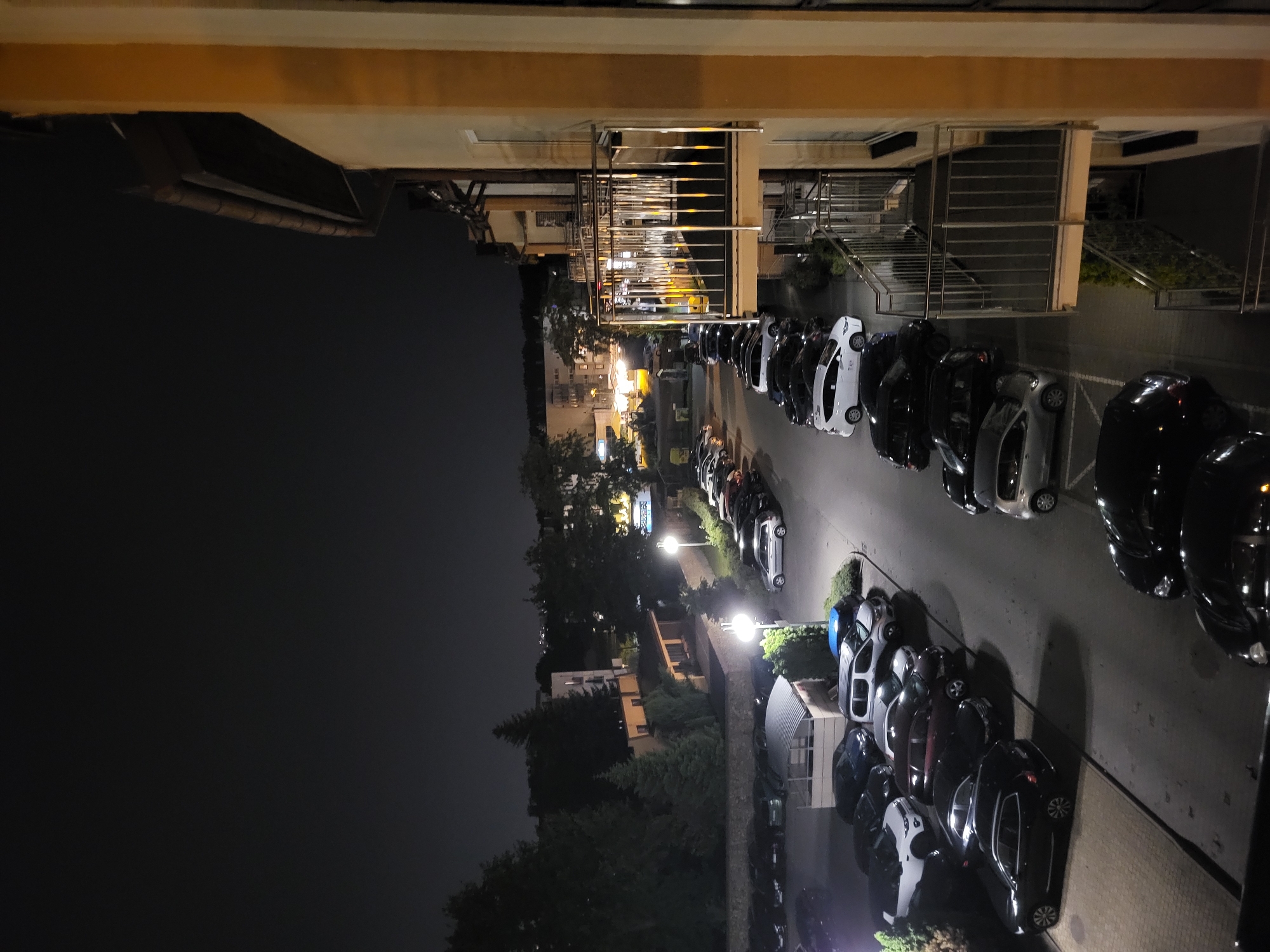 Here is a description of the image:
The image shows a night view of a parking area located in an urban environment. The area is filled with parked cars in a row, illuminated by streetlights. In the background, buildings and trees are visible under the night sky. The photograph appears to have been taken from an elevated position, possibly from a building overlooking the parking area.