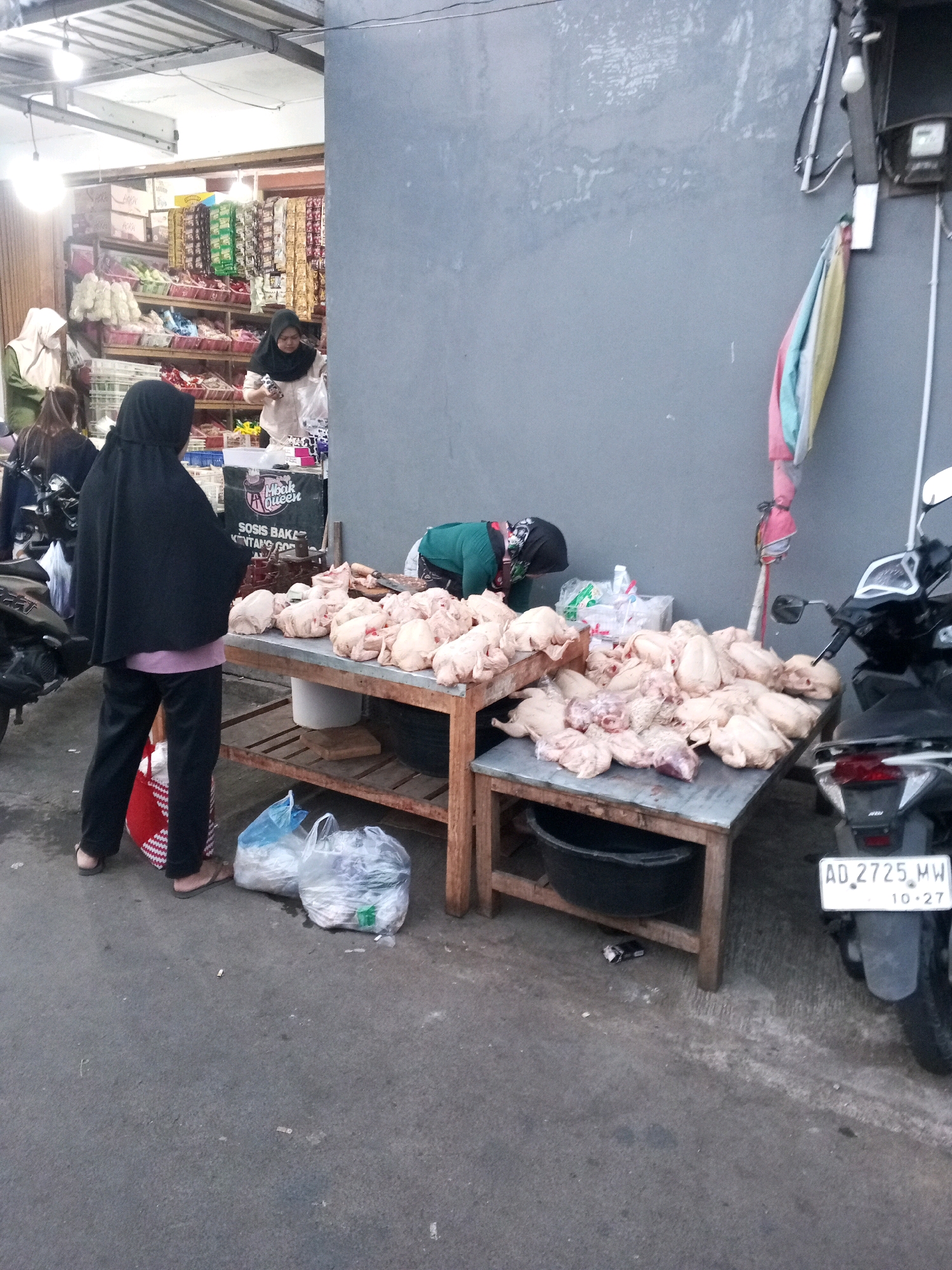 In Gentan, Indonesia, a street market scene unfolds with two women at a stall selling raw chickens. One woman, wearing a black hijab and a pink shirt, stands facing away from the camera, holding a red and white striped bag. The other woman, in a green headscarf, is bent over the table, attending to the chickens. A third woman in a black hijab is visible behind them, further into a shop. The chickens are displayed on a wooden table, with some packed in plastic bags. A motorcycle is parked to the right of the stall, and the ground is paved. The scene appears to be outdoors, with a building wall providing a backdrop.