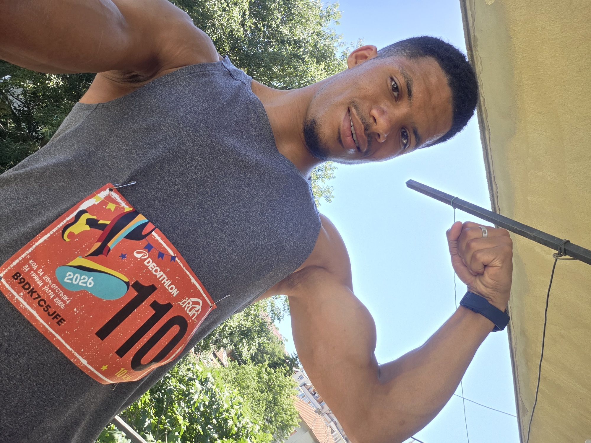 A man of color is taking a selfie outdoors, flexing his bicep. He is wearing a gray tank top with a race bib pinned to it. The bib is orange and has the number 110 prominently displayed. Text on the bib appears to be in Bulgarian. He is also wearing a dark fitness tracker on his left wrist and a ring on his right index finger. The background consists of a clear blue sky, green trees, and buildings. Based on the text on the bib, which includes "Varna 2026" and "10KM", it is likely that the image was taken in Varna, Bulgaria, during or in preparation for a 10-kilometer race in 2026.