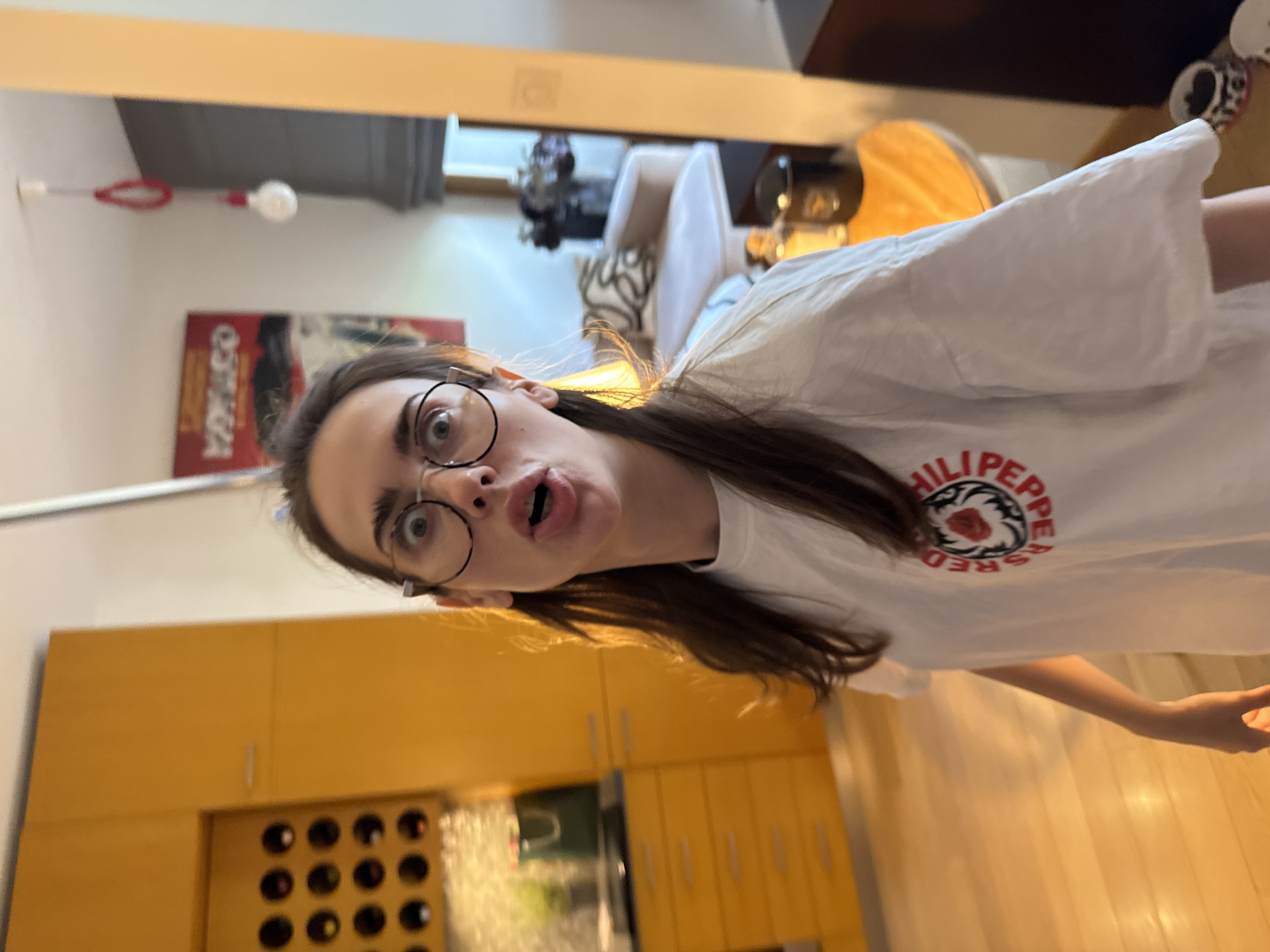 A person with glasses is facing the camera with a surprised expression. The person is wearing a white T-shirt with a red and black graphic design. The background includes a kitchen with wooden cabinets, a wine rack, and a poster on the wall. The location is an interior space in the city of Unknown, Unknown.