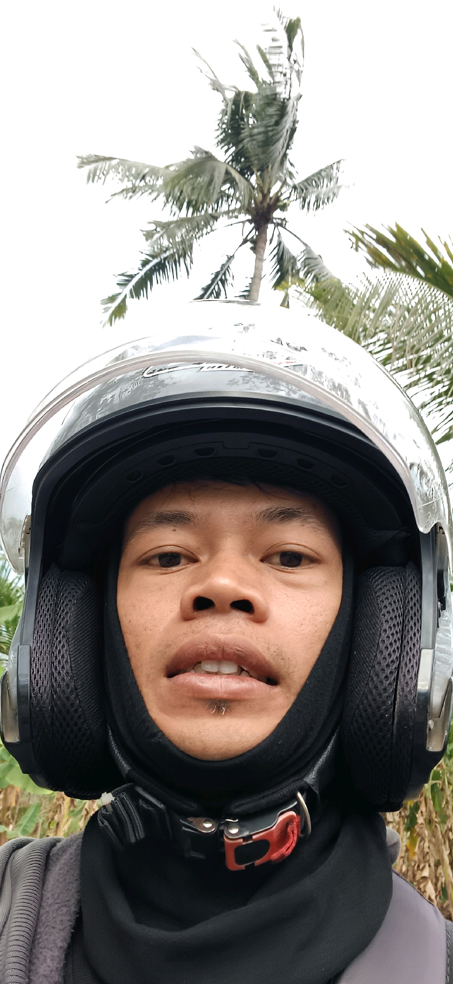 A person wearing a black motorcycle helmet with a clear visor is looking directly at the camera. The helmet has mesh ear coverings. The person has dark eyes and dark hair. There is a small silver ring on their lower lip. They are wearing a black neck gaiter and a grey garment. In the background, a palm tree is visible against a bright sky. The image appears to be taken outdoors, possibly in an area with vegetation. The location is identified as Badran, Indonesia.