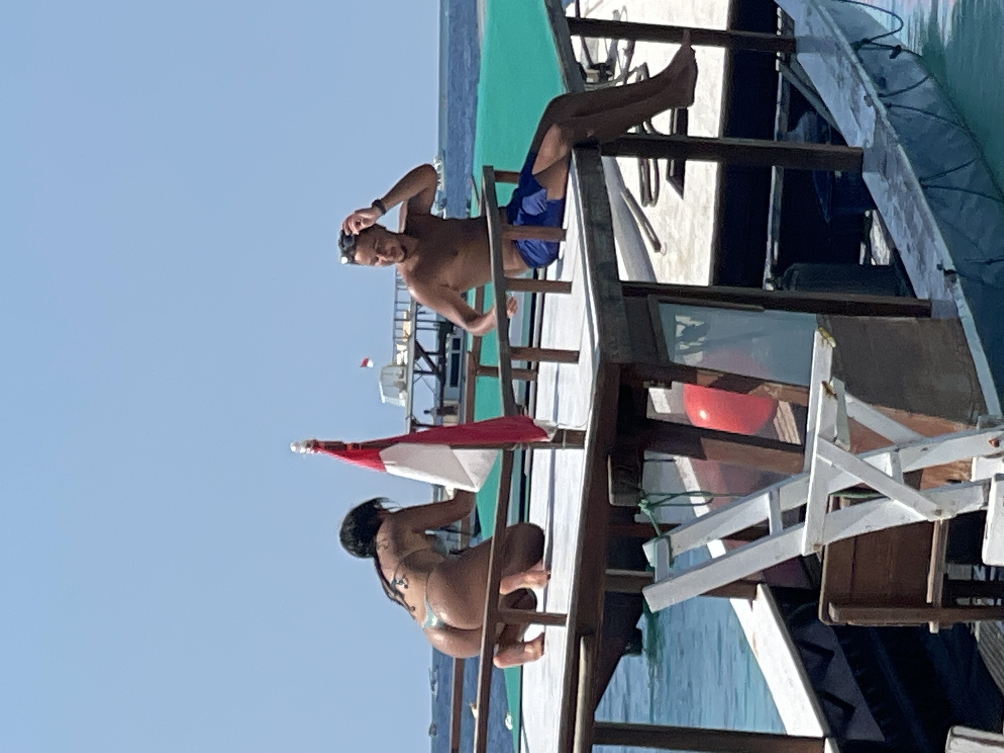 On a bright, sunny day, two individuals are on the deck of a boat. In the foreground, a woman with dark hair is kneeling on the deck, facing away from the camera. She is wearing a light-colored bikini. A red and white flag is attached to a pole behind her. In the middle ground, a man with short dark hair is sitting on the edge of the deck, to the right of the woman. He is shirtless and wearing blue swim trunks. He has one hand on his head, and sunglasses are perched on his hair. Both individuals appear relaxed. The boat is in the water, with the turquoise ocean visible around it. In the background, other boats and structures can be seen on the water. The sky is clear and blue.