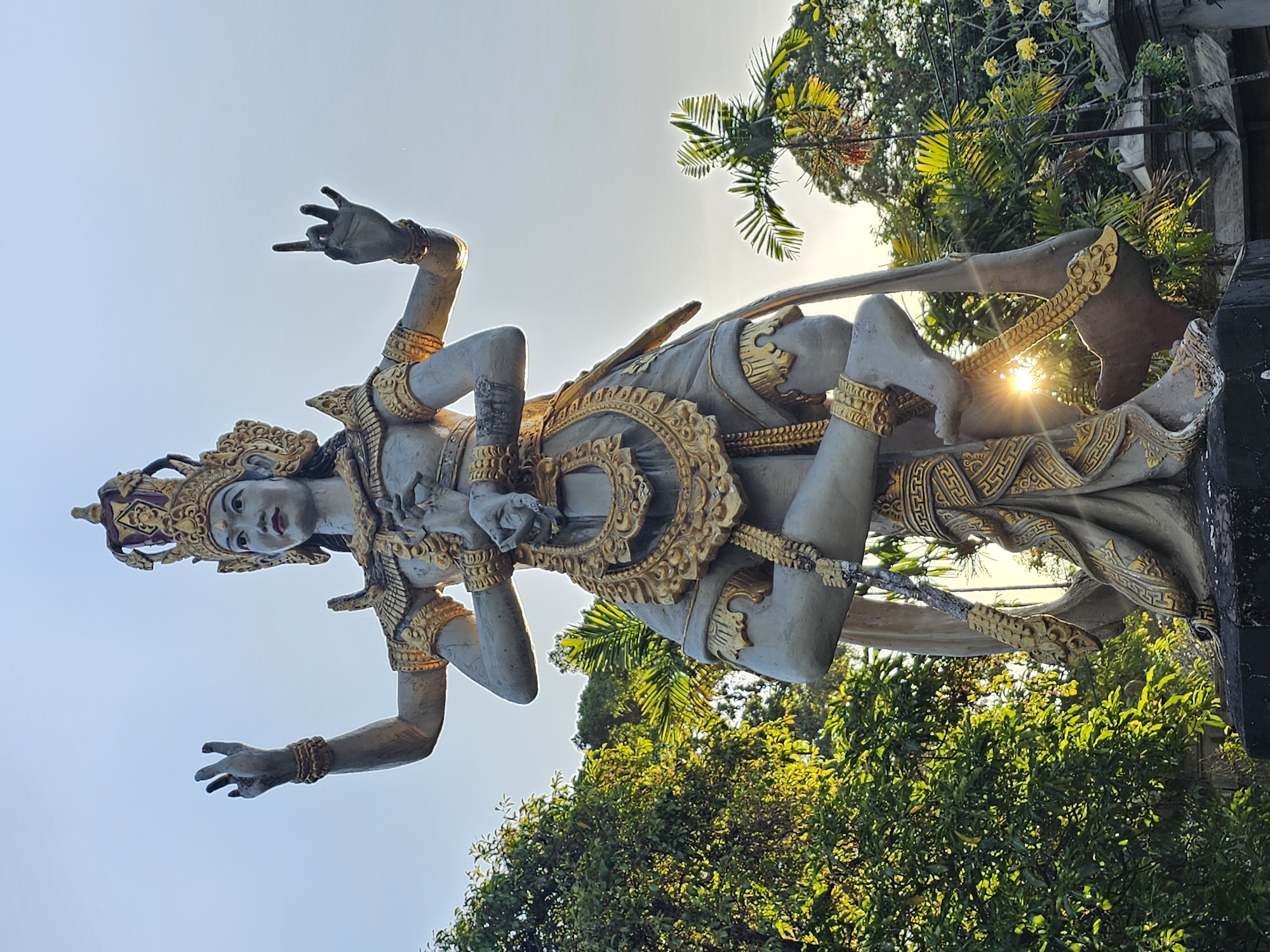 A full-body, low-angle shot captures a large, gray and gold statue of a multi-limbed deity against a bright, hazy sky. The statue, depicting a figure with a crown, intricate jewelry, and flowing garments, is positioned in a dynamic pose with legs crossed and arms raised and bent. The sun shines through the statue's lower body, creating a lens flare effect. Lush green trees and palm fronds are visible in the background, partially obscuring the sky. The statue appears to be located in an outdoor setting, possibly a temple or garden, with glimpses of architectural elements, including a stone balustrade and what might be a part of a wall, to the right. The overall atmosphere suggests a warm, possibly tropical, environment. The statue's intricate detailing and the sunlight create a visually striking and somewhat ethereal impression. The statue is likely situated in Denpasar, Indonesia, known for its rich cultural heritage and numerous religious sites.