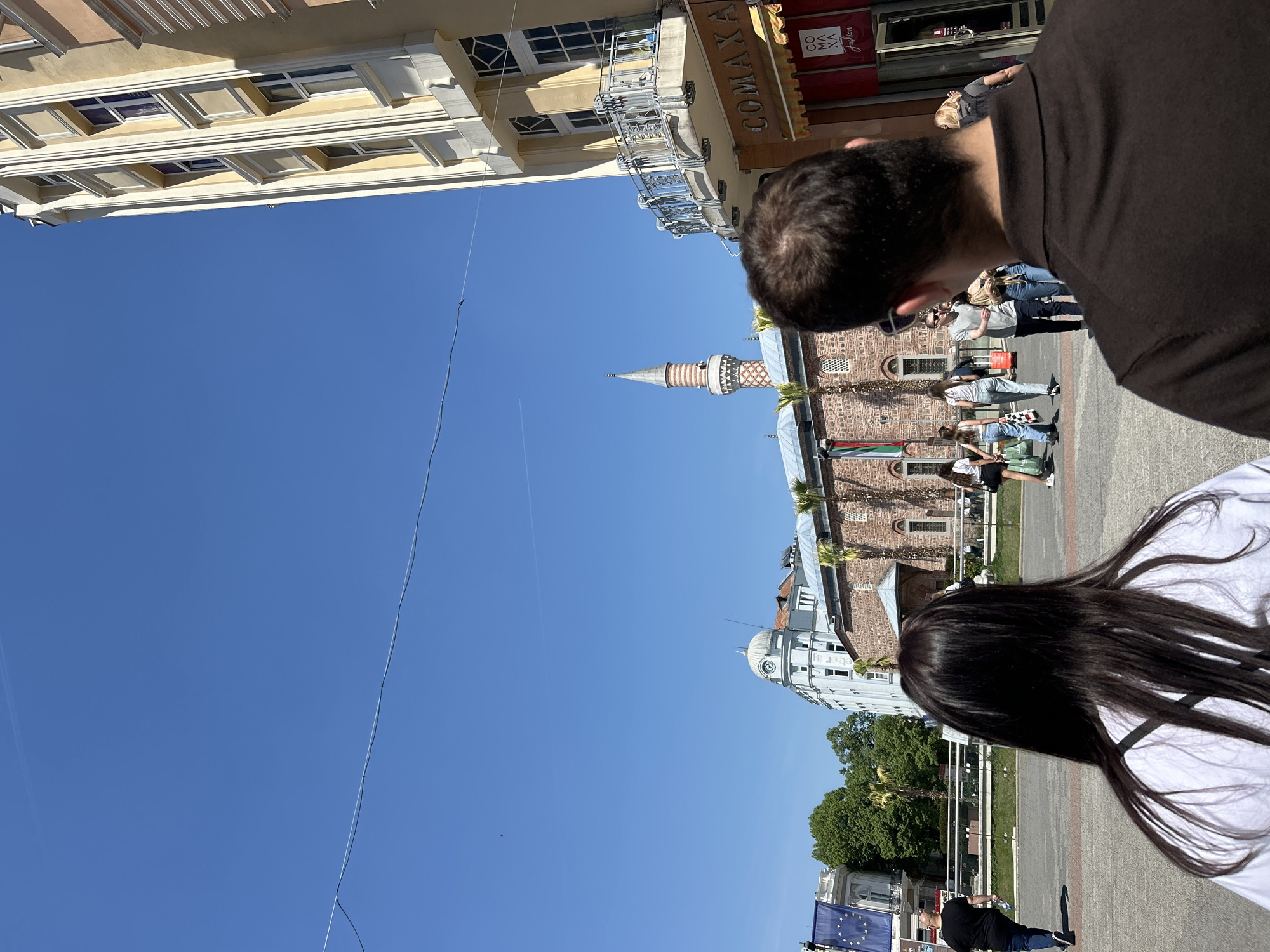 The image captures a scene in Plovdiv, Bulgaria, with a clear blue sky overhead. The perspective is from ground level, looking upwards. The Jumaya Mosque, a historic building with a distinctive minaret, is prominent in the center of the frame. Adjacent to the mosque is a building, possibly administrative, constructed in a light color with classic European architectural details. A building with clock tower is also visible on the bottom of the frame, slightly off-center. Several people can be seen in the middle-ground, interacting and moving around the plaza area. The figures are walking on a paved surface. The presence of the EU flag hints at the city's integration with European affairs. In the foreground, the backs of two people's heads and shoulders are visible. A man in a dark shirt is on the right, and a woman with long black hair in a white shirt is on the bottom right. A building facade on the top of the frame, possibly a storefront, has a sign written in Cyrillic. Scaffolding is visible on the building. Overhead wires cut across the sky.