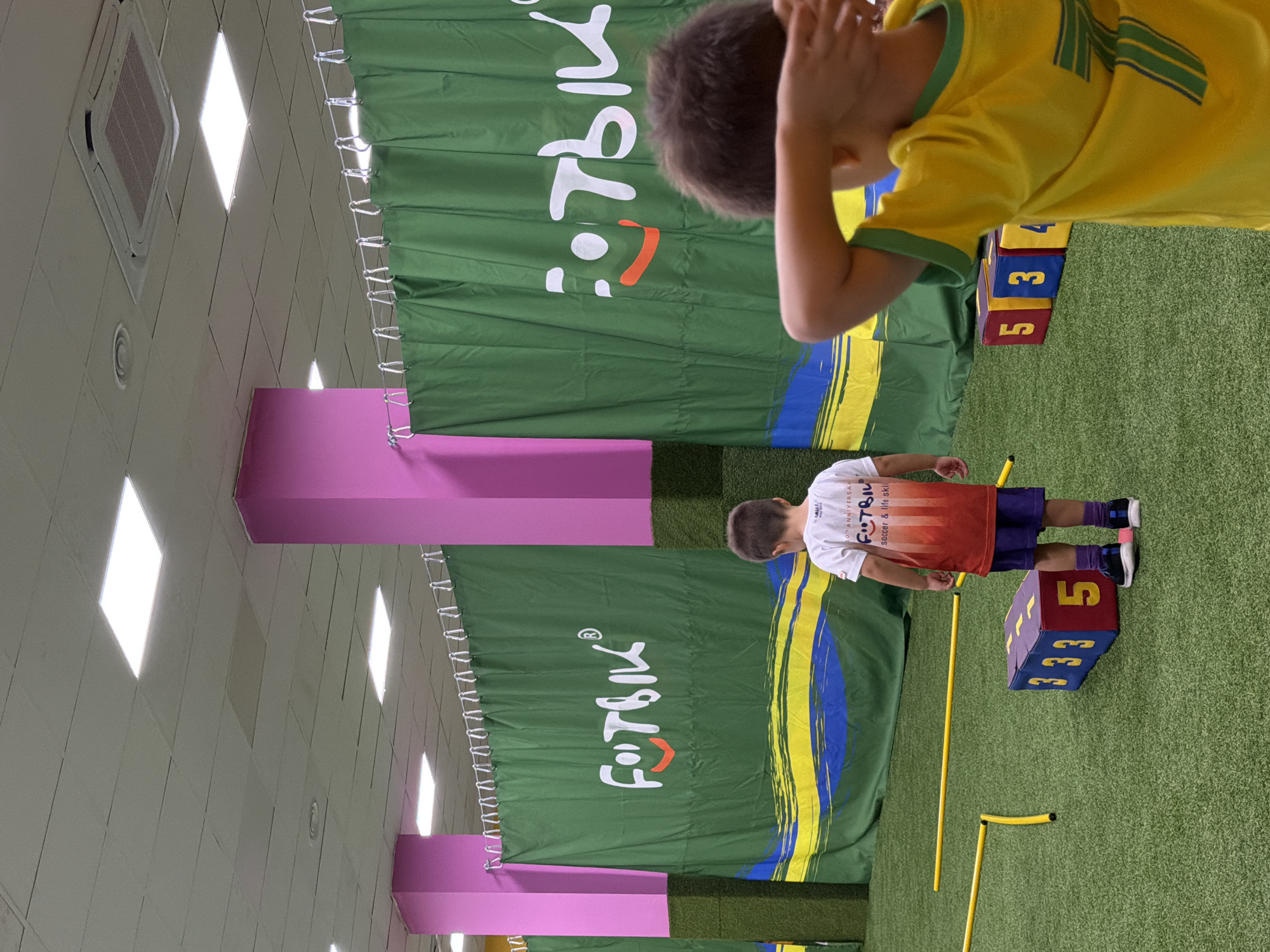 In this image, a young boy wearing a yellow and green soccer jersey is seen from the back, his hands near his head, suggesting he might be engaged in a game or exercise. He is positioned in an indoor setting with a green, artificial turf floor. Behind him, a large green banner with a white logo that reads "FOOTBIL" and a smiling face is displayed. A second, similar banner is visible further back in the scene. There are also large pink rectangular structures in the background.
In the foreground, another young boy is standing with his back to the camera, wearing a white t-shirt with an orange and red gradient design and the "FOOTBIL" logo. He is wearing purple shorts and dark athletic shoes. He is standing next to a cube with the number "5" on one side and "3" repeated on another. Yellow poles are set up on the turf floor, indicating a training or obstacle course. The lighting suggests an indoor environment, possibly a sports facility or gymnasium.
The text visible includes "FOOTBIL" on the banners and t-shirt, and numbers on the cube. The overall impression is one of a children's sports activity or training session.