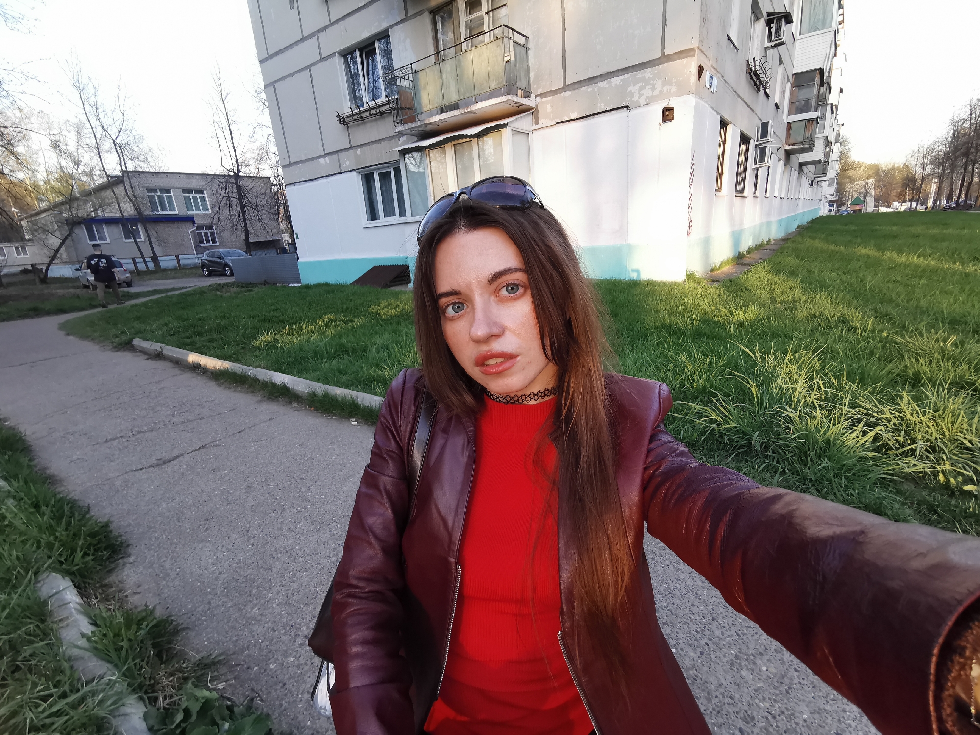 A young woman stands on a sidewalk in an urban area. She is wearing a red shirt and a maroon leather jacket, and has sunglasses on her head. A choker is visible around her neck. To her left is a building with multiple floors and balconies, partially painted white. To her right, there is a grassy area. In the background, another building is visible, along with trees and a sidewalk where a man is walking. A car is parked next to the building in the background.