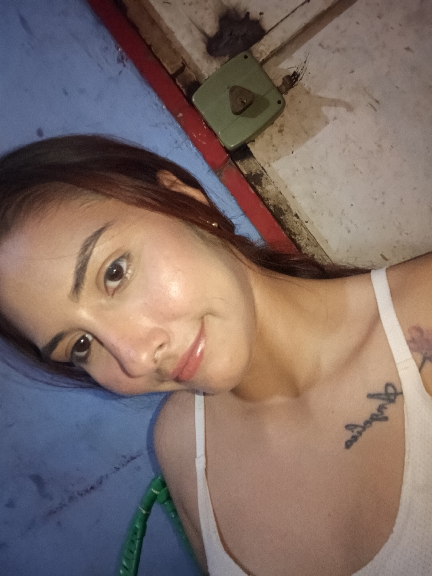 A woman is shown in a selfie. She has brown hair, light skin, and is wearing a white tank top. There is a tattoo on her upper chest. A green plastic chair is partially visible in the bottom left. The background is a blue wall next to a floor and wall corner area with a green and metal latch. The location, Juan Griego, Venezuela, is not visually confirmed.
