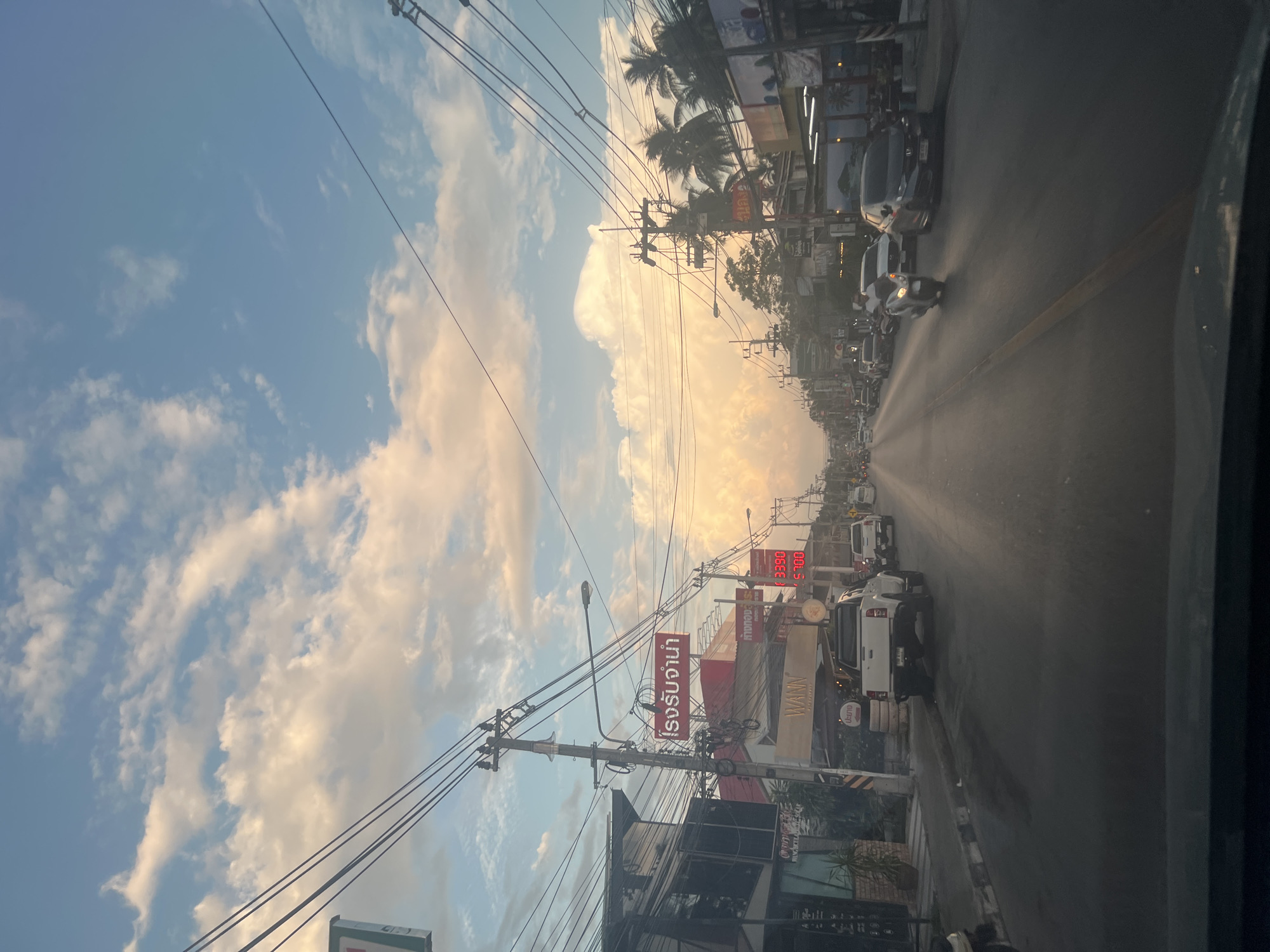The image captures a street scene in the Ko Samui District of Thailand, likely during the late afternoon or early evening, given the warm, diffused light. The sky is a pale blue with scattered clouds, some of which are illuminated with a golden hue. Overhead, a complex network of power lines crisscrosses the frame, supported by utility poles lining the street.
On the left side of the road, several buildings are visible, including a shop with a dark facade and large windows, and a prominent red sign with Thai script. Another building, seemingly a business with "WANN" displayed on its awning, is also present. A white pickup truck is parked on the side of the road, facing away from the viewer.
The road itself is paved and appears to be a main thoroughfare with multiple lanes. Several vehicles, including cars and a scooter with a rider, are traveling on the road, mostly heading into the distance. Palm trees and other tropical vegetation are visible on the right side of the street, interspersed with more buildings and signs, some of which display illuminated numbers. The overall impression is of a bustling commercial area in a tropical setting.