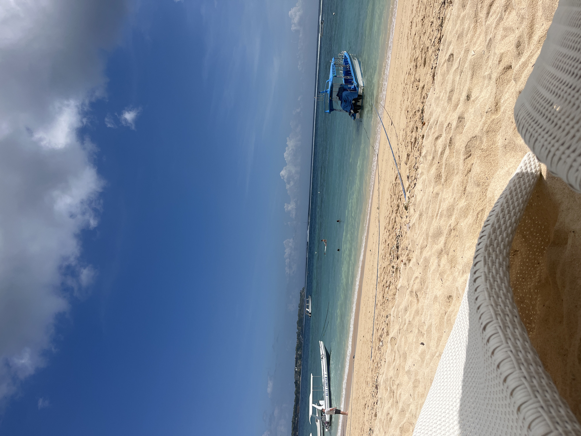 The image captures a view from a beach in Nusa Penida, Indonesia. In the foreground, a white woven lounge chair is partially visible, casting a shadow on the golden sand. The beach stretches towards the turquoise ocean, where gentle waves are breaking. Several boats are anchored in the shallow water. One boat, predominantly blue with a white hull, is closest to the shore and appears to be a passenger boat. Further out, a white boat with a canopy is visible, and another smaller boat is further in the distance. A person is standing on the beach near the white boat with the canopy. In the water, several individuals are swimming or wading. The sky is a clear blue with scattered white clouds. The overall scene depicts a tranquil tropical beach setting.