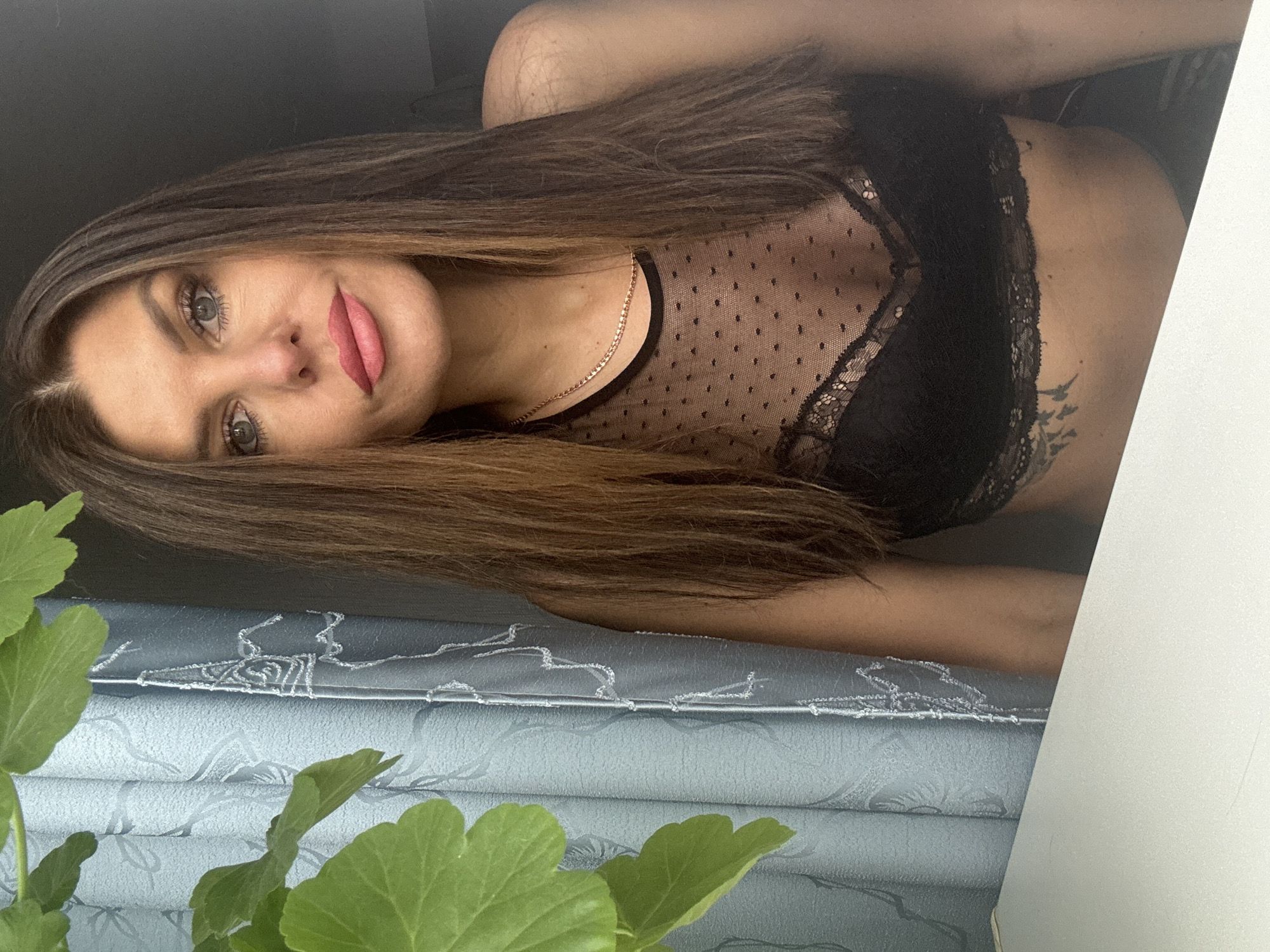 The image features an adult female captured in an indoor setting, positioned beside a window with light blue patterned curtains. A green leafy plant is out of focus in the immediate left foreground. The woman has long, straight brown hair with subtle highlights, parted near the center, falling over her shoulders and chest. She has blue-grey eyes, wears noticeable eye makeup, and bright pink lipstick. Her expression is neutral with a direct gaze toward the viewer. She is wearing a sheer black mesh top with small polka dots over a black lace bralette. A thin, rose gold-colored chain necklace is visible around her neck. A dark, intricate tattoo is partially visible on her left side, just above her waistline. A flat, white surface, likely a desk or windowsill, occupies the lower foreground. The background behind the woman's right side is dark and indistinct. There are no discernible elements within the image that allow for confirmation or identification of the location as Dubovaya, Russia.