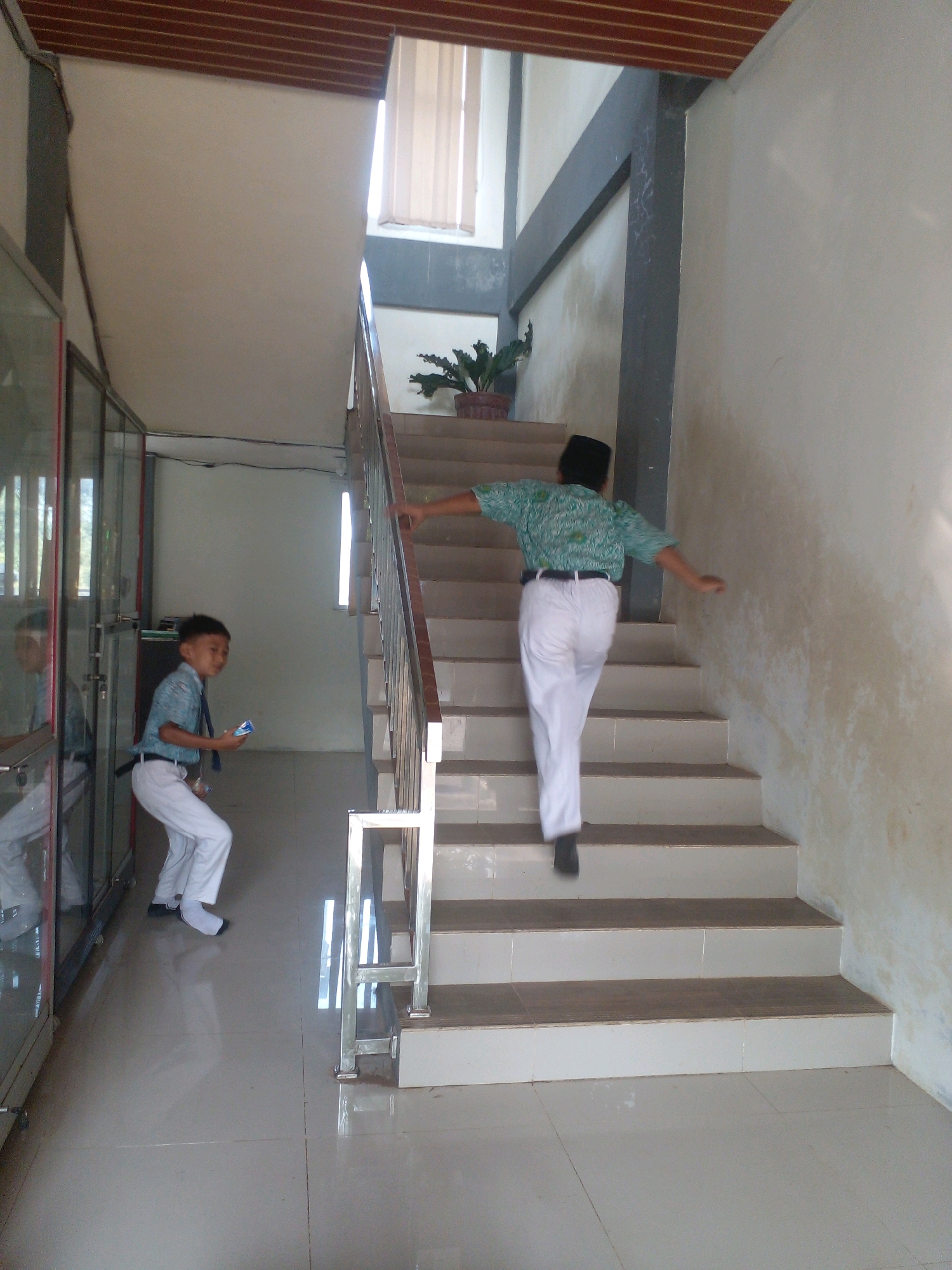 The image shows a indoor staircase with two young boys in school uniforms. The boy on the left, wearing a light blue shirt and white pants, is standing on the tiled floor and holding a small blue object. He appears to be looking to his right. The boy on the right, wearing a green patterned shirt and white pants, is walking up the stairs, with his back to the camera. He is wearing a dark skullcap. The staircase has metal railings and the walls are painted a light color, with some discoloration. There is a potted plant on the landing of the stairs. To the left of the staircase, there are glass-fronted cabinets or doors. The ceiling has wooden slats. The scene appears to be within a school or educational facility.